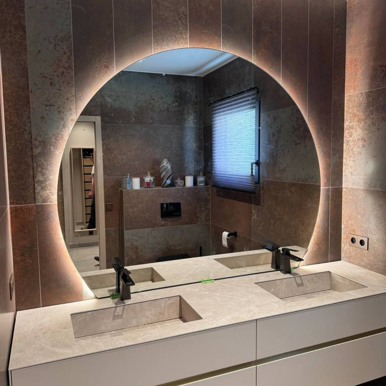 Salle de bains moderne avec miroir rond rétroéclairé au-dessus d'une double vasque. Murs carrelés marron et fenêtre visibles.
