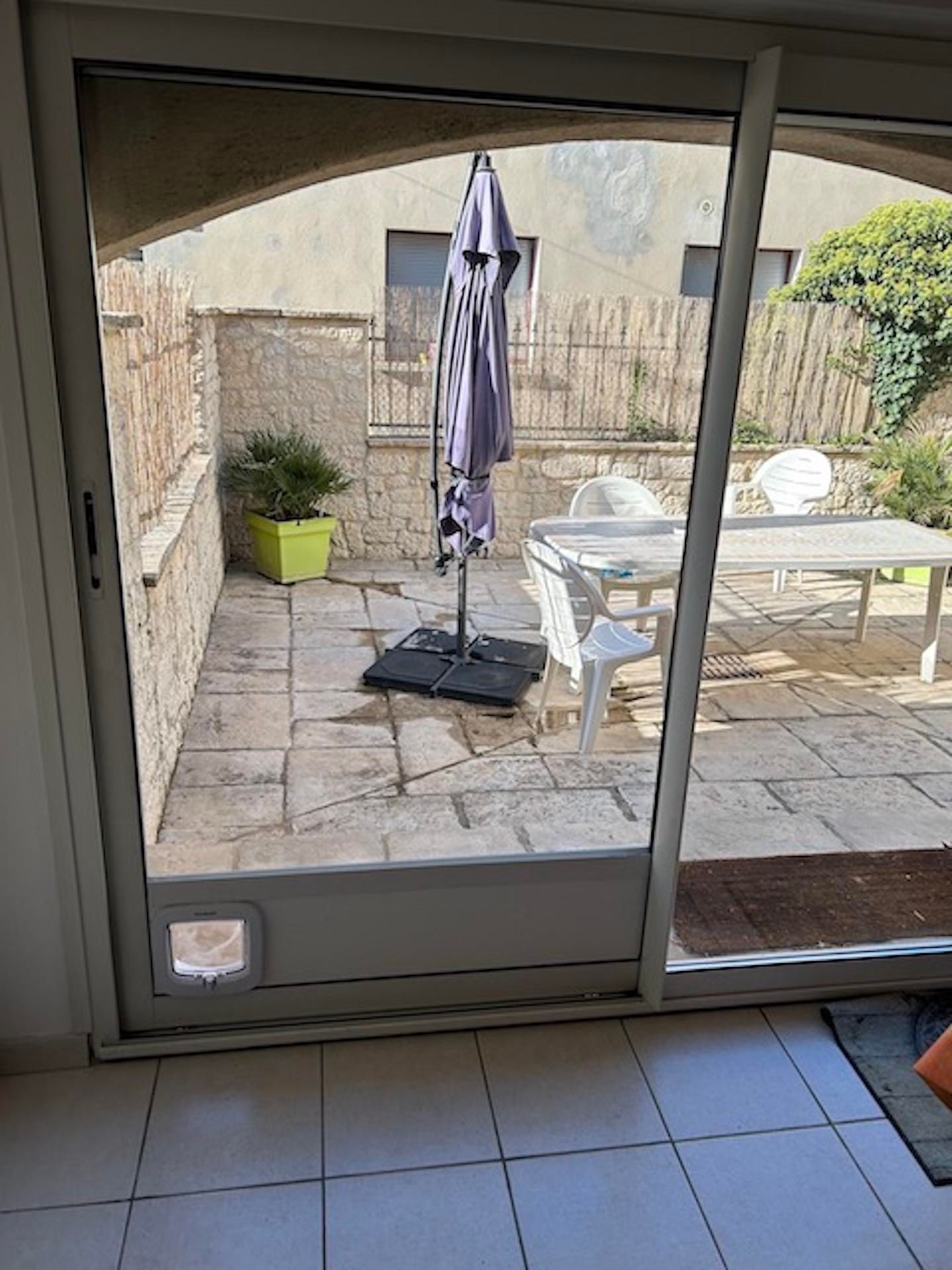 Une porte coulissante en verre avec chatière ouvre sur une terrasse équipée d'une table, de chaises et d'un parasol fermé.