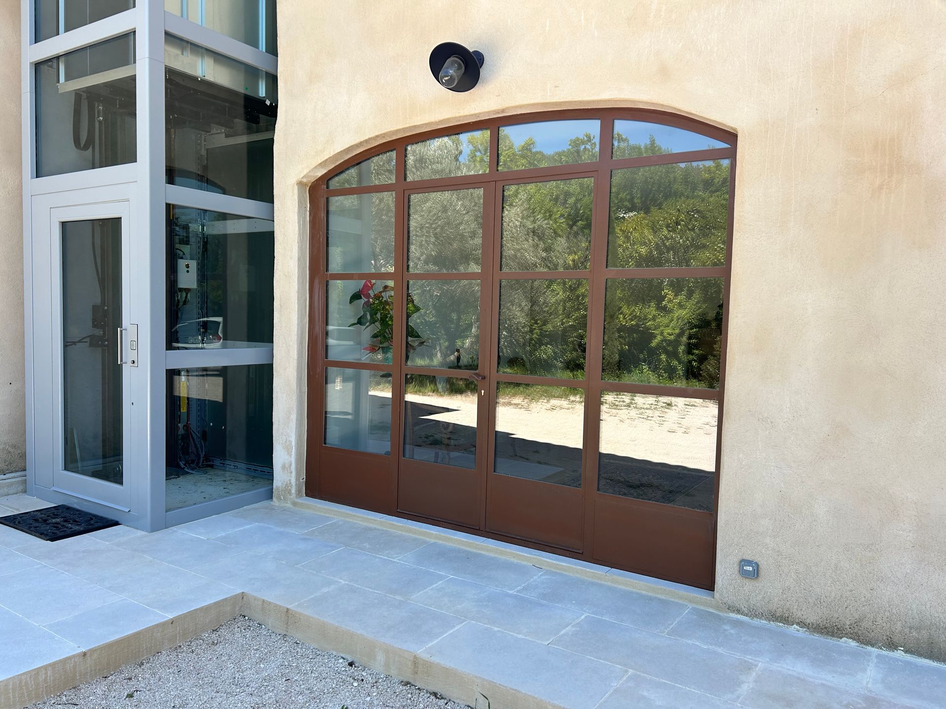 Vue extérieure d'un bâtiment avec un ascenseur en verre et des portes métalliques brunes cintrées reflétant des arbres.
