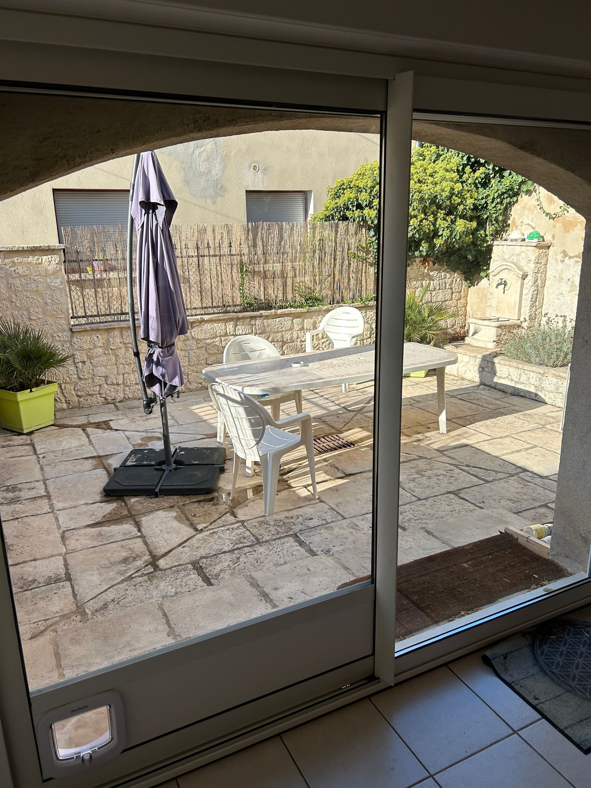 La porte coulissante en verre s'ouvre sur un patio avec table, chaises, parasol et chatière.