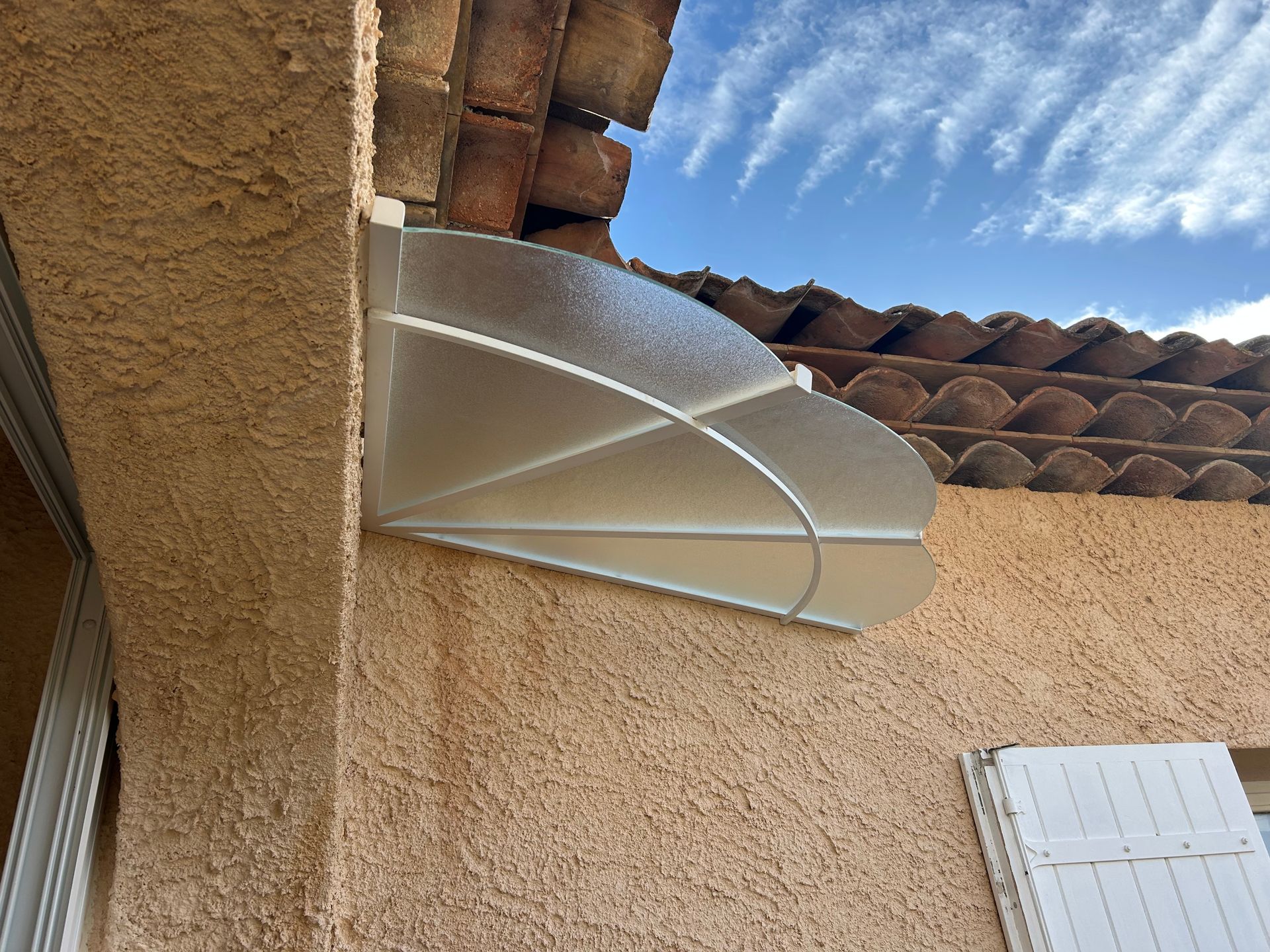 Auvent transparent et arqué monté sur un mur en stuc sous un toit en tuiles, contre un ciel bleu.