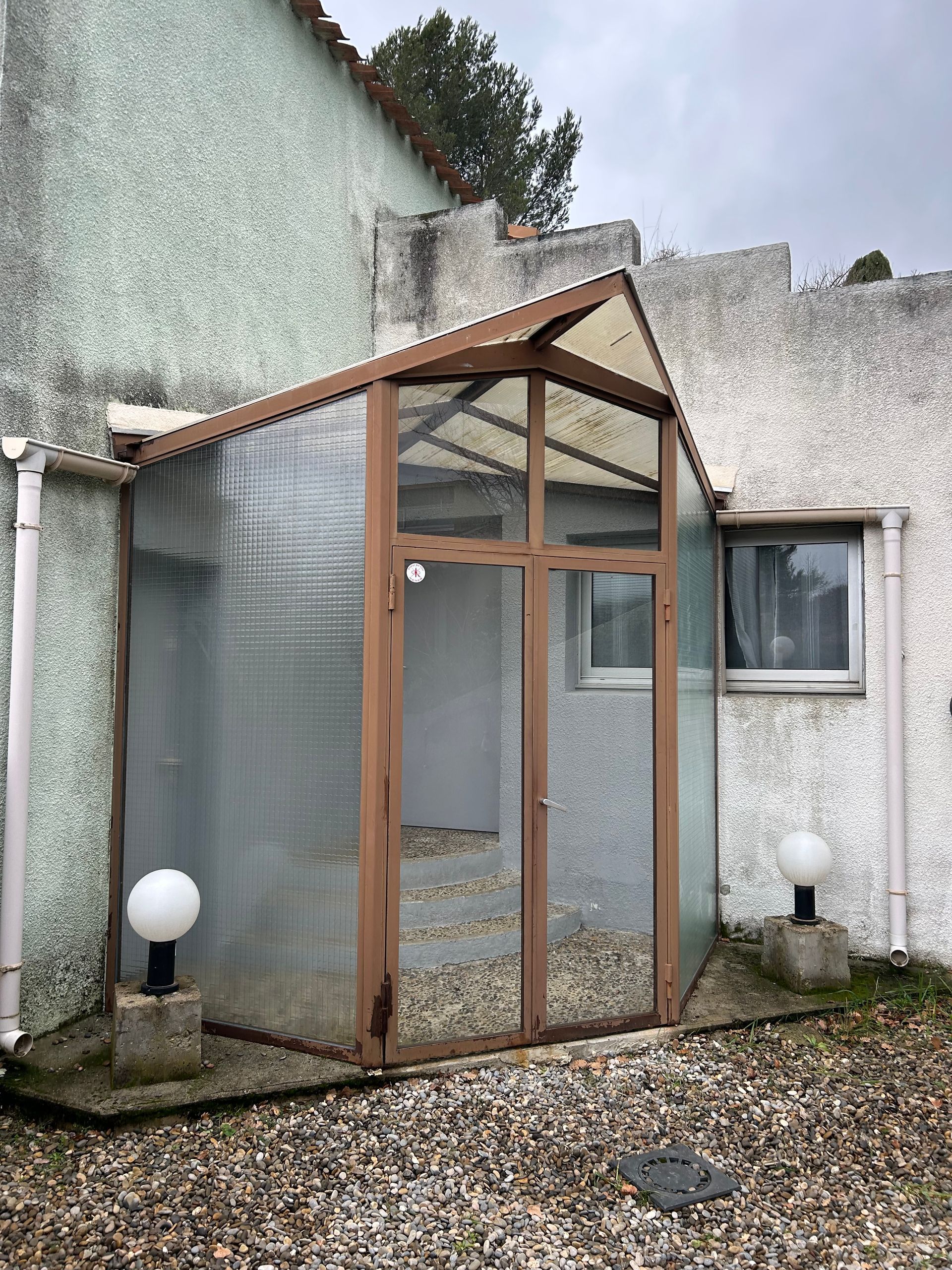 Abri d'entrée aux parois en plastique transparent et au cadre marron, menant à la porte et à l'escalier d'un bâtiment.