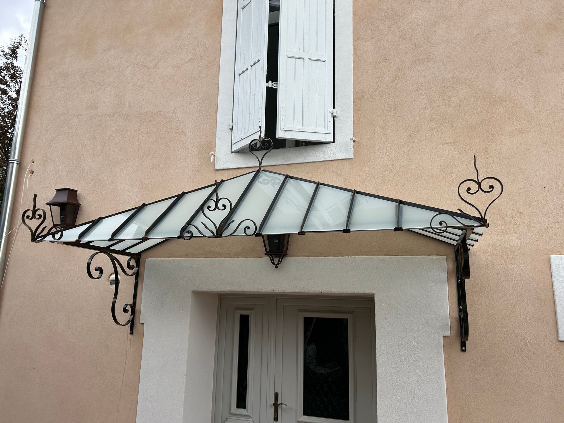 Auvent en verre et fer forgé au-dessus d'une porte blanche sur un bâtiment rose clair, avec une fenêtre au-dessus.