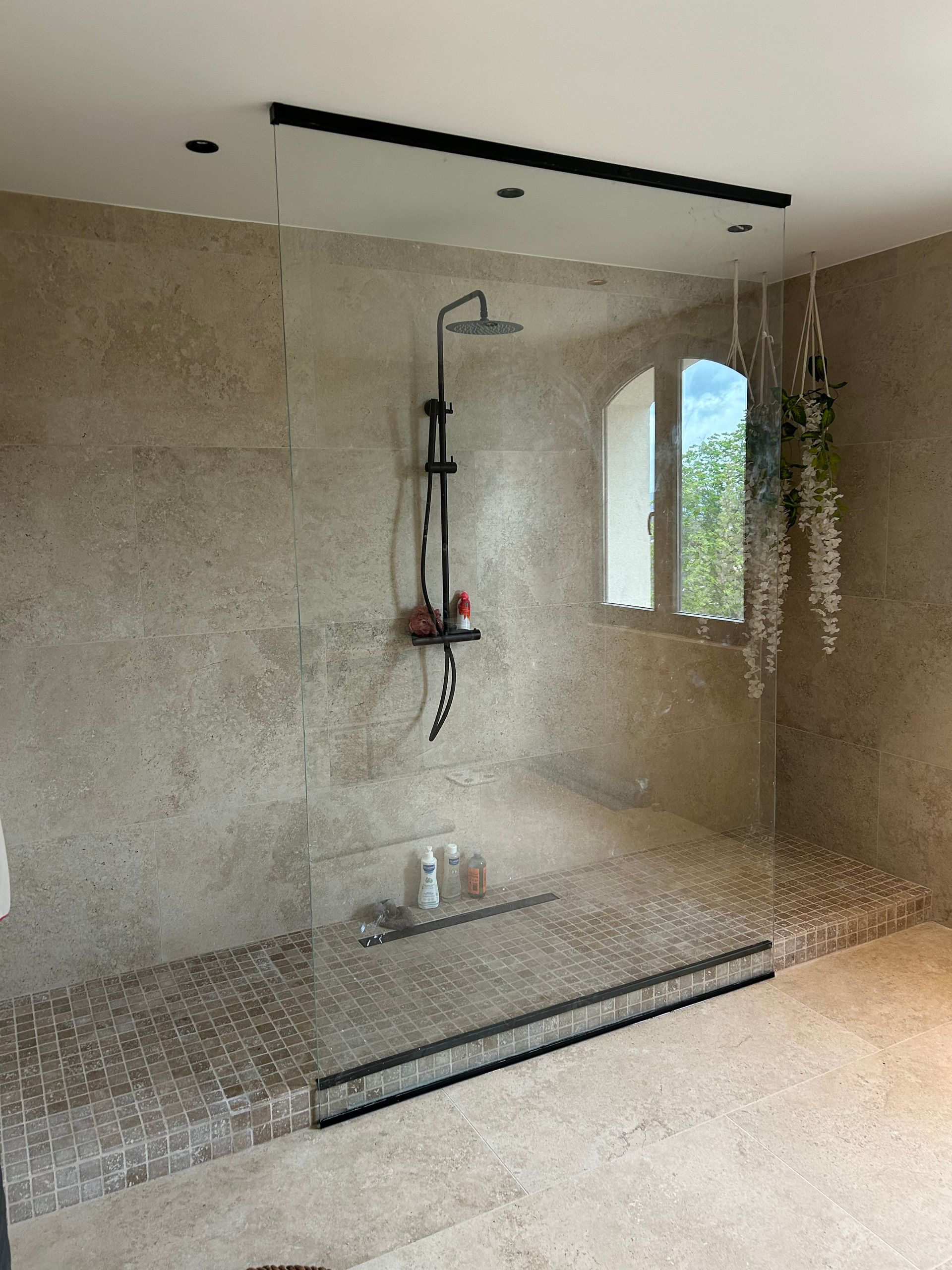Douche avec sol en galets, cabine en verre, robinetterie noire et fenêtre avec plantes.