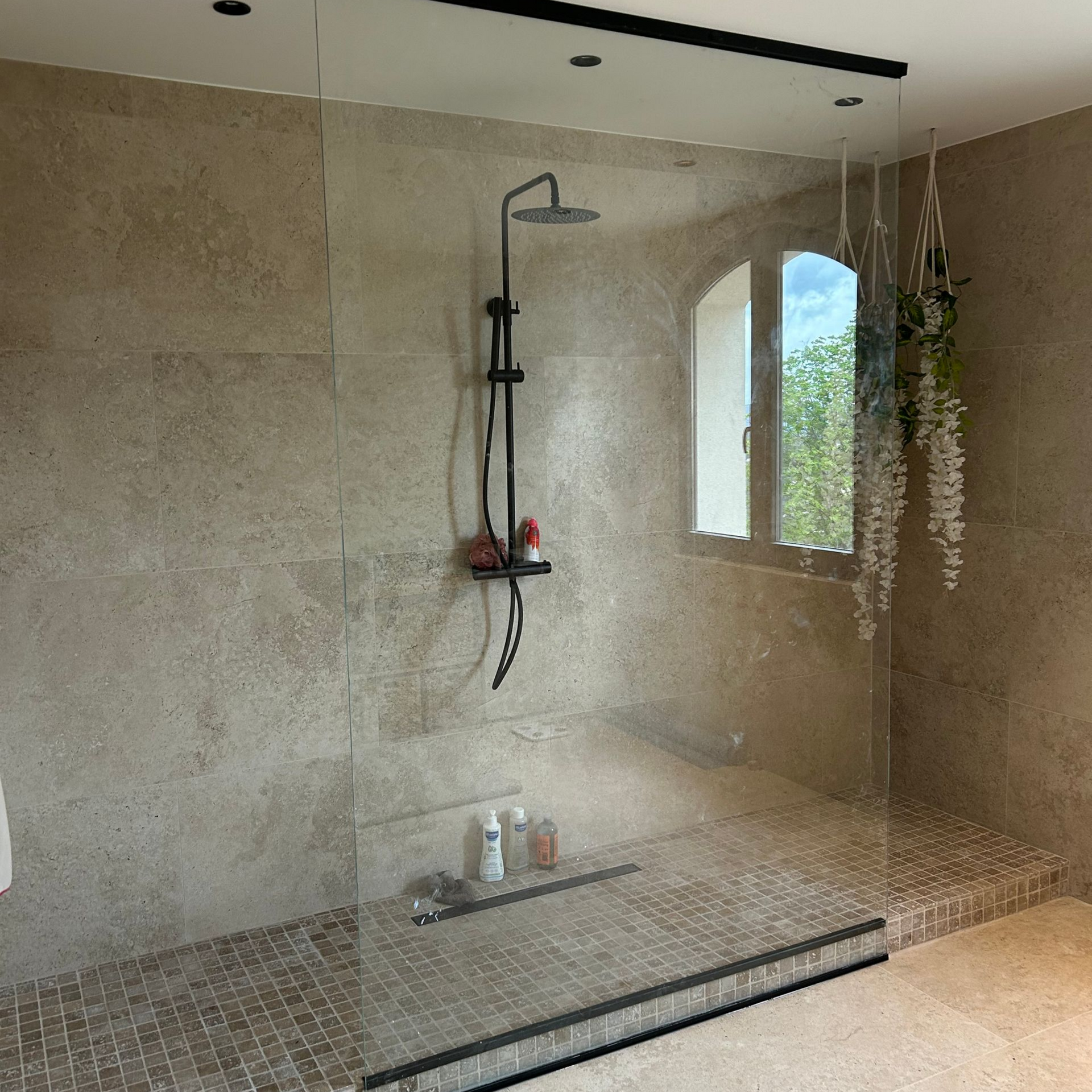 Douche avec paroi en verre transparent, pommeau noir et carrelage en pierre.