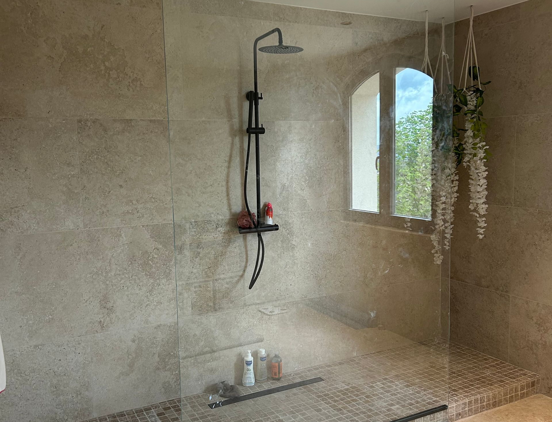 Douche avec murs en carrelage de pierre, robinetterie de douche noire, fenêtre avec vue extérieure.