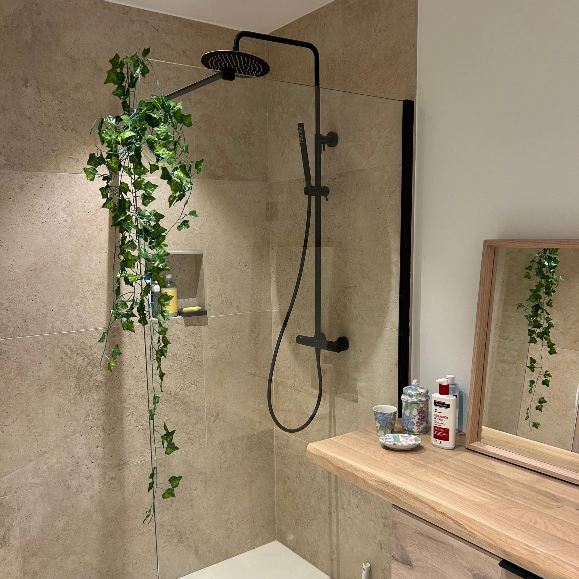 Douche avec robinetterie noire, murs en pierre et verdure suspendue; vanité en bois avec miroir.