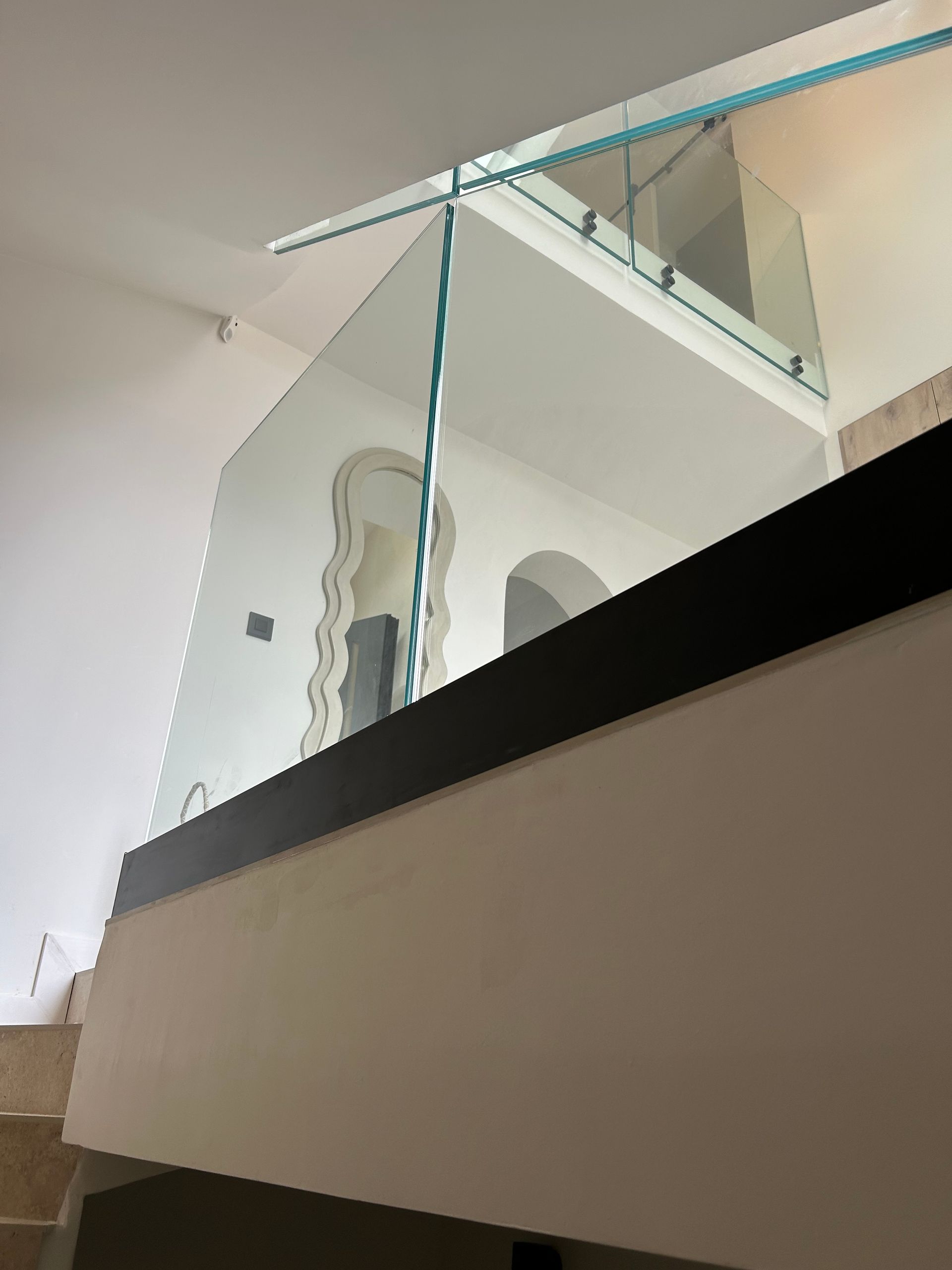 Garde-corps en verre sur un balcon intérieur avec murs blancs, porte cintrée et luminaires.