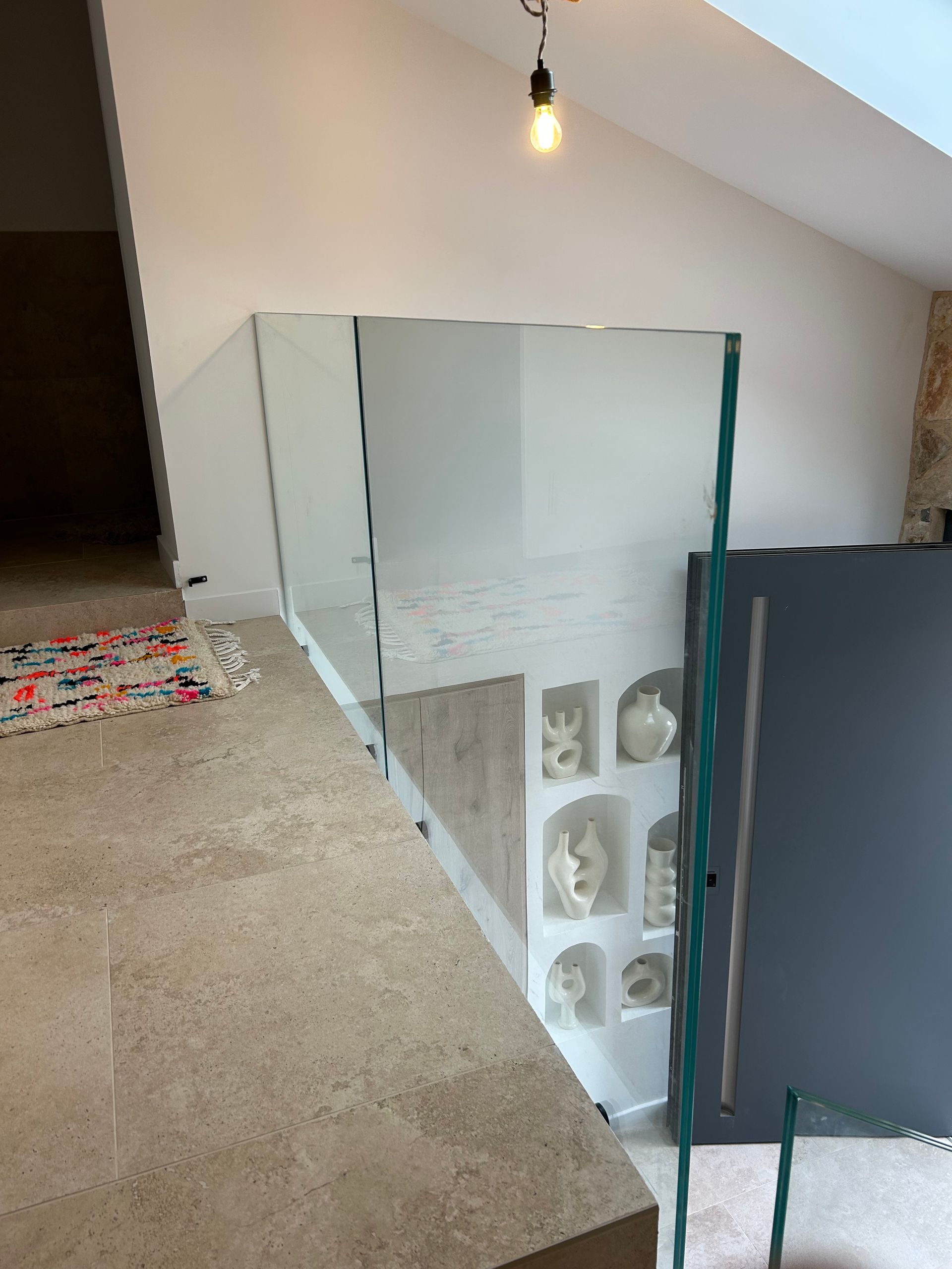 Rampe d'escalier en verre dans une maison moderne. Sol en pierre beige, murs blancs.