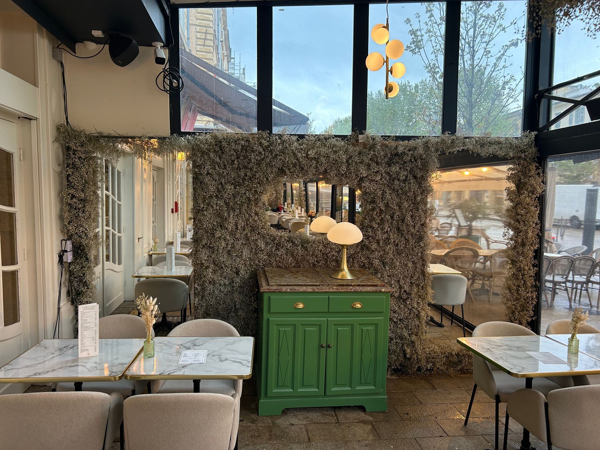 Intérieur d'un café avec tables en marbre, armoire verte et mur recouvert de fleurs séchées.