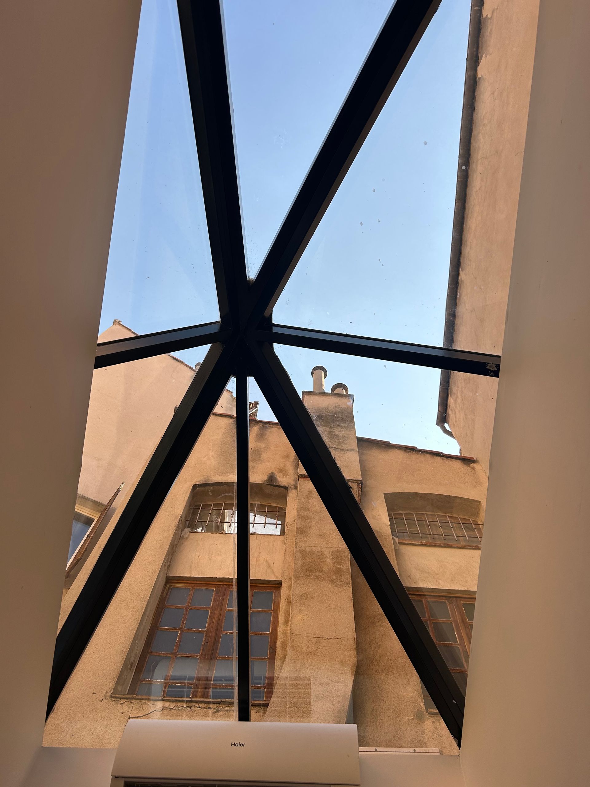 Vue depuis le puits de lumière sur les bâtiments extérieurs et le ciel bleu, avec des poutres transversales sombres.