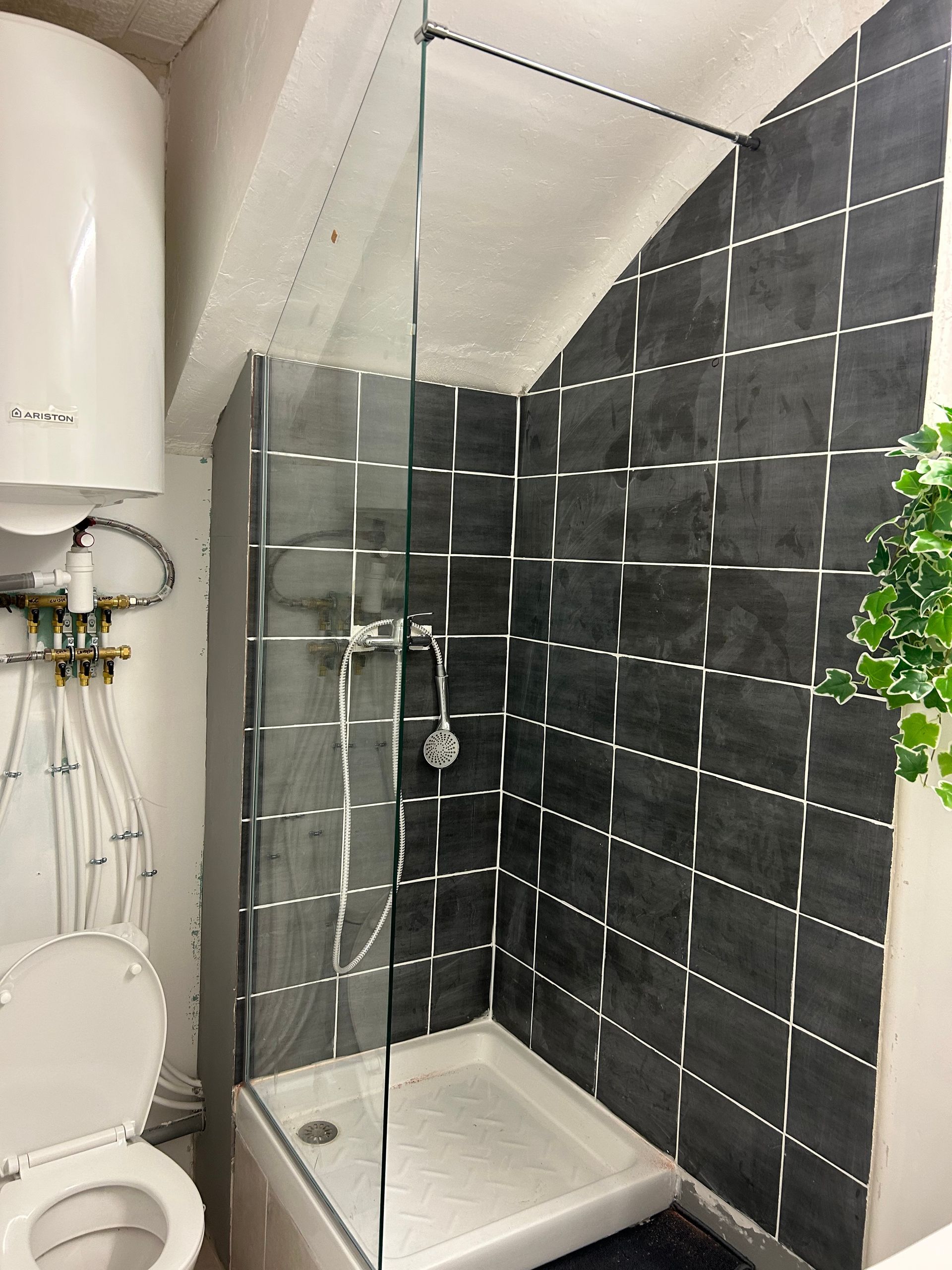 Petite salle de bain avec toilettes, douche et chauffe-eau. La douche est carrelée de noir et dotée d'une paroi vitrée.