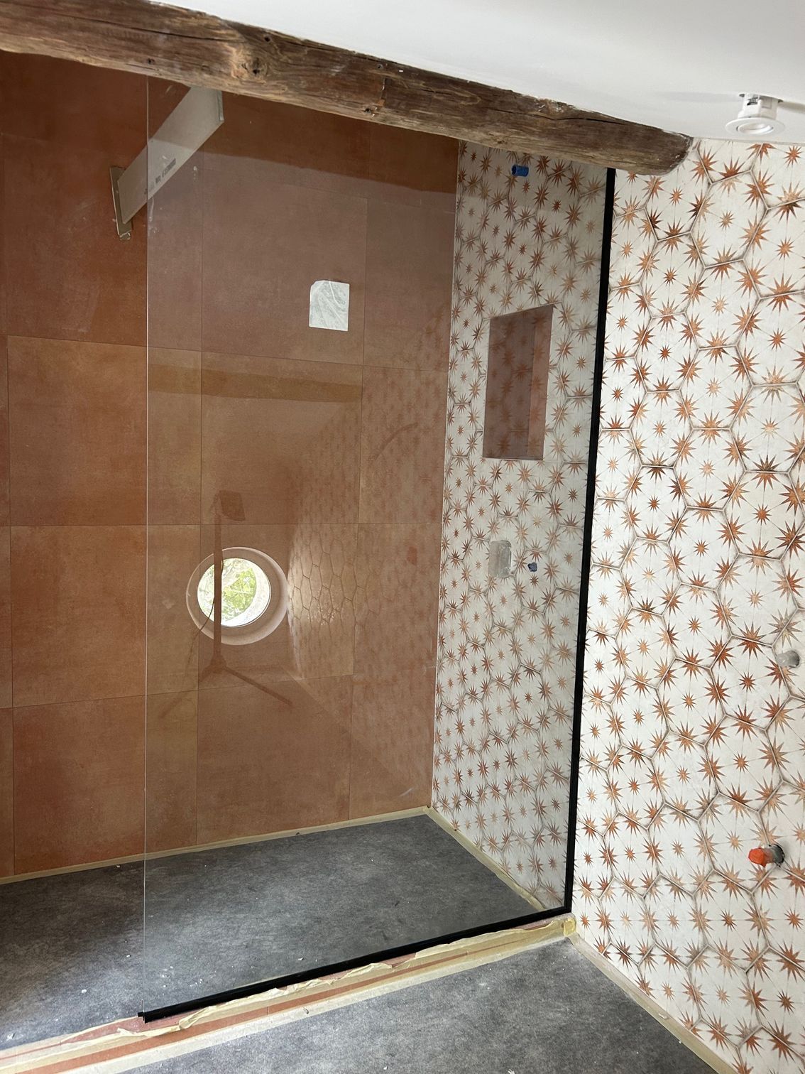 Douche avec porte vitrée, carrelage marron, mur décoratif, sol foncé et poutre en bois.