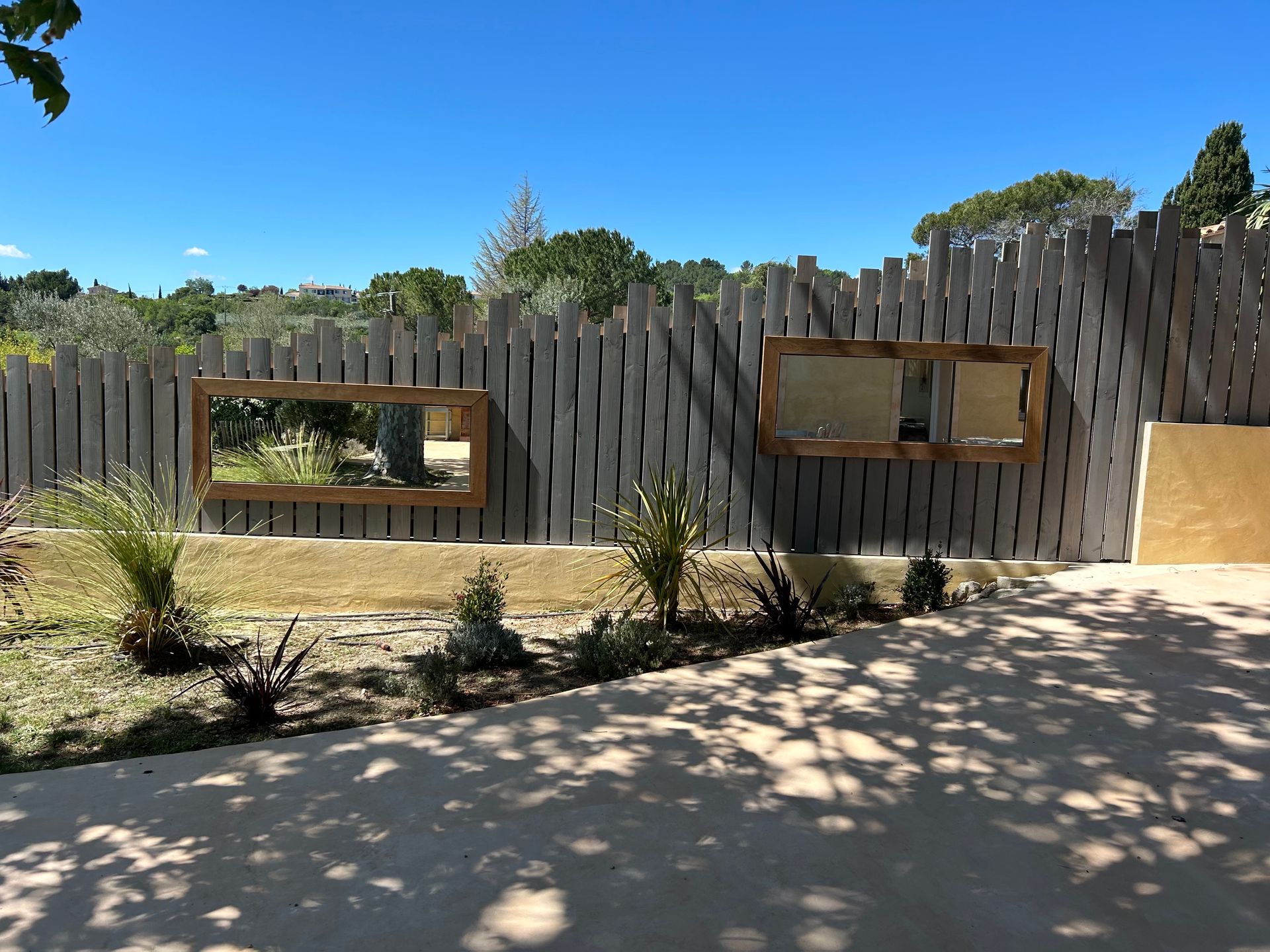 Clôture en béton avec deux miroirs encadrés, journée ensoleillée, petites plantes devant.