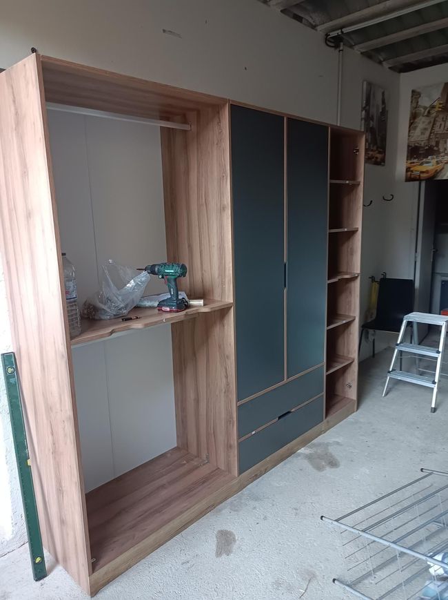 Armoire encastrée inachevée dans une pièce, avec des moulures en bois et des étagères sombres, outils à proximité.