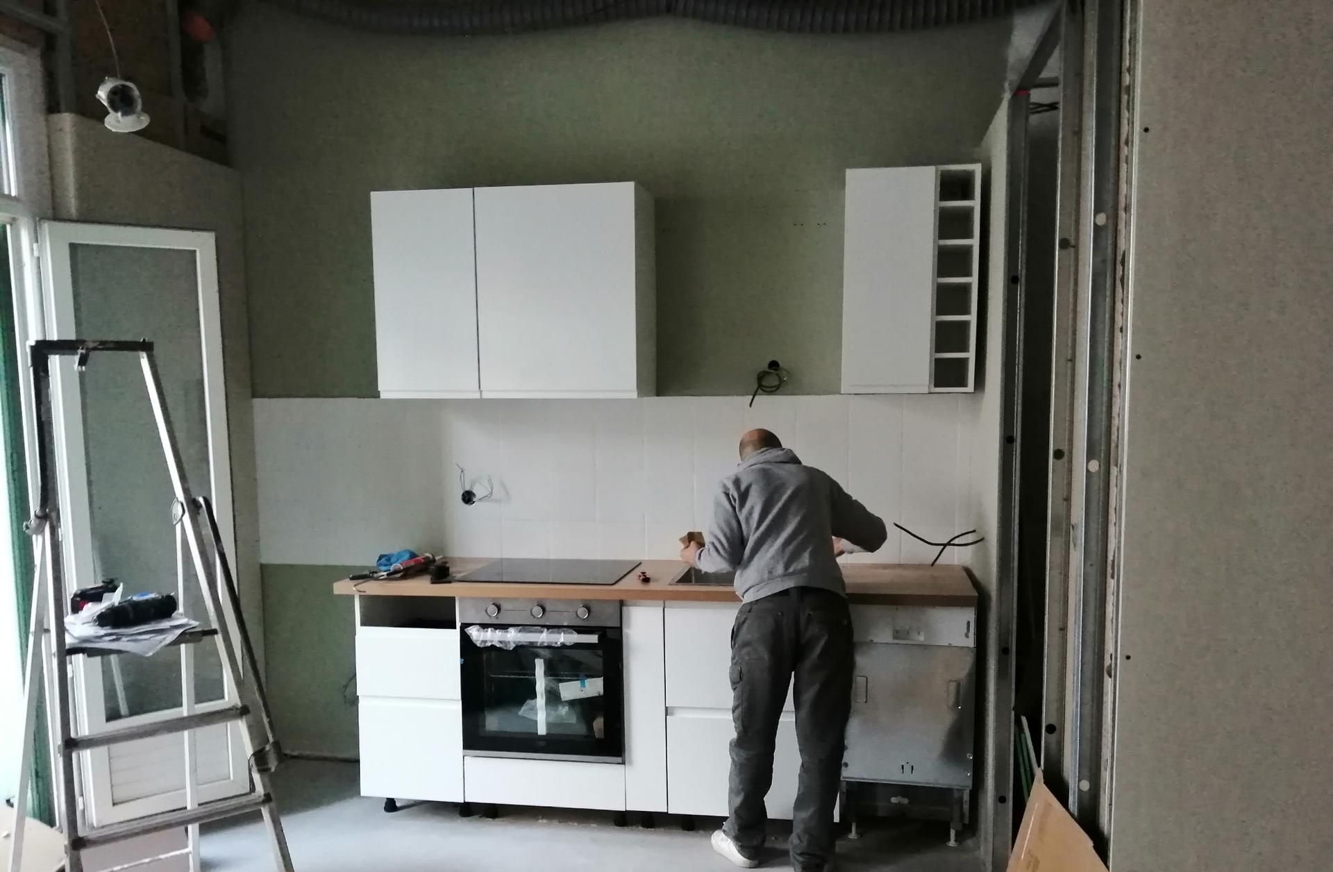 Une personne installe des armoires de cuisine et un comptoir blancs dans une pièce non finie.