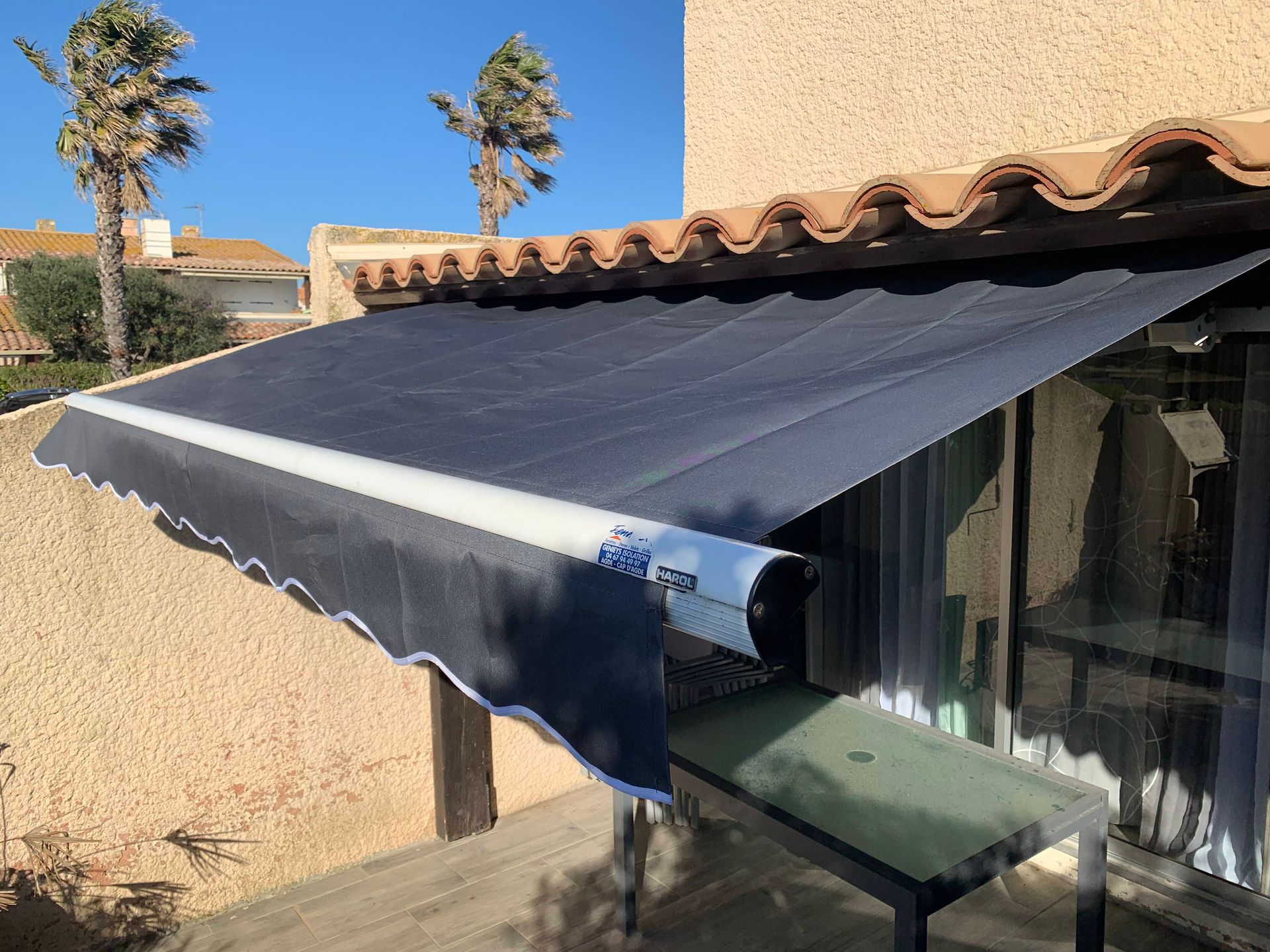 Store banne rétractable noir au-dessus d'une terrasse jouxtant une maison, avec portes vitrées et toit en tuiles.