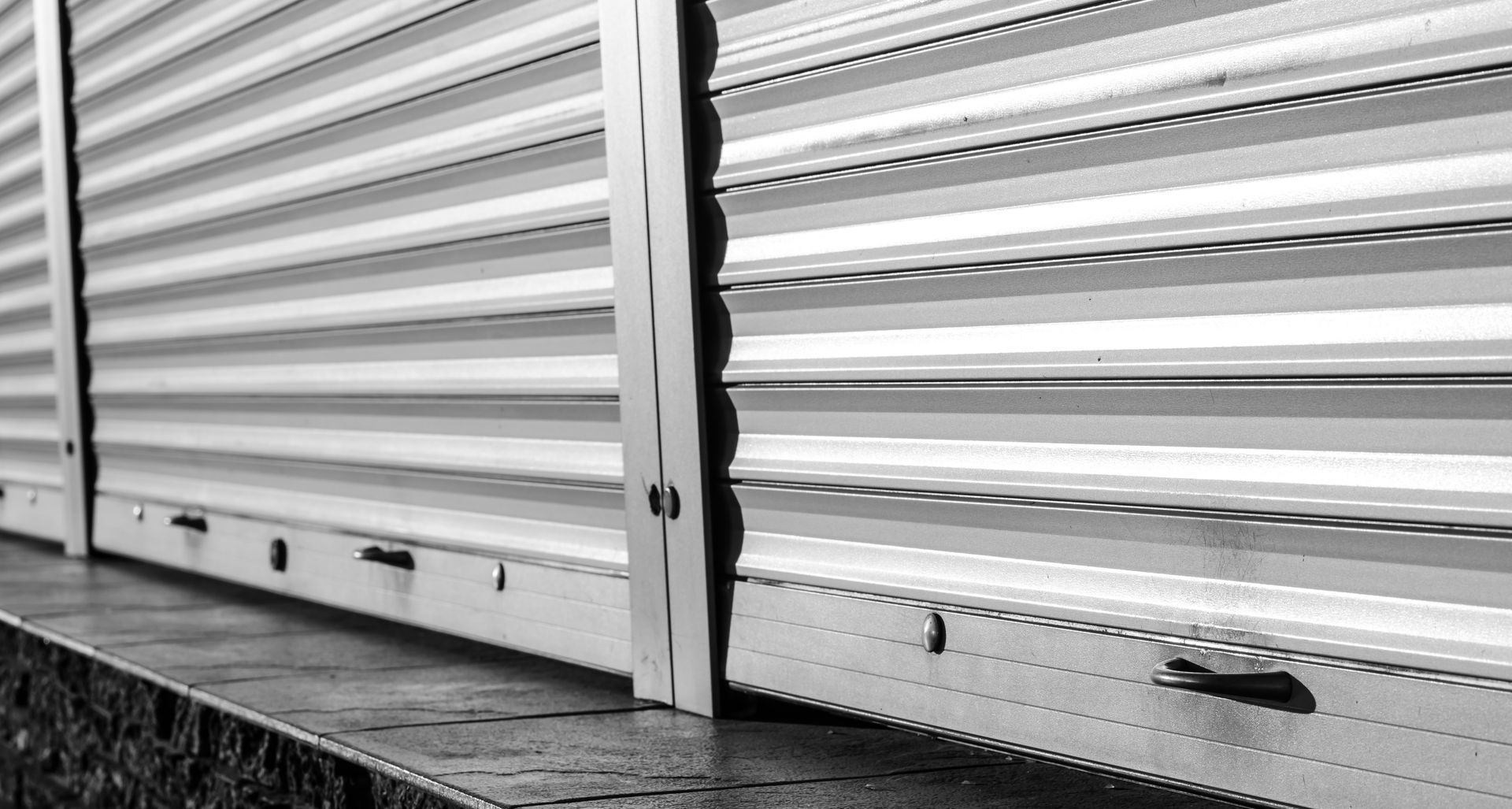 Volets roulants métalliques blancs le long d'un trottoir, vus en gros plan en noir et blanc.