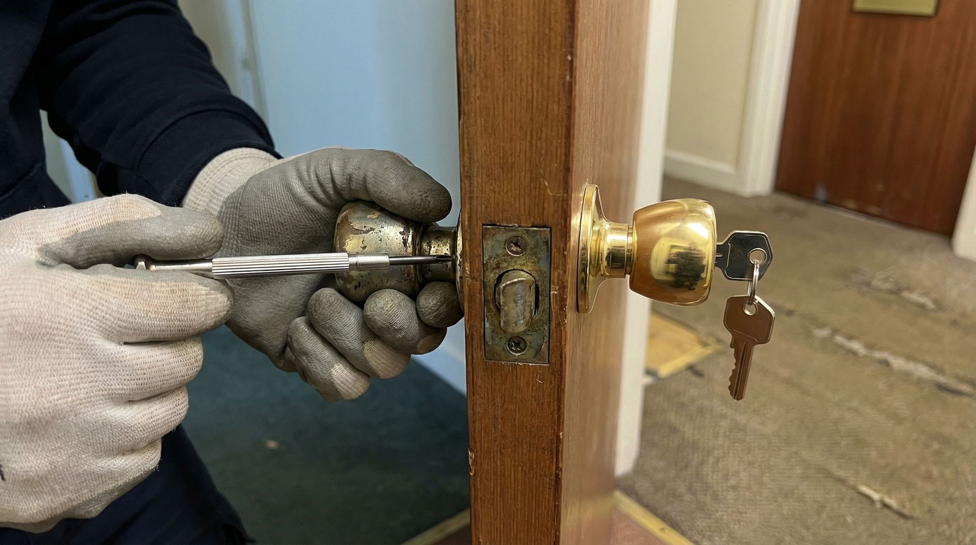 Des mains gantées utilisent un tournevis pour ajuster une serrure de porte dorée sur une porte en bois.