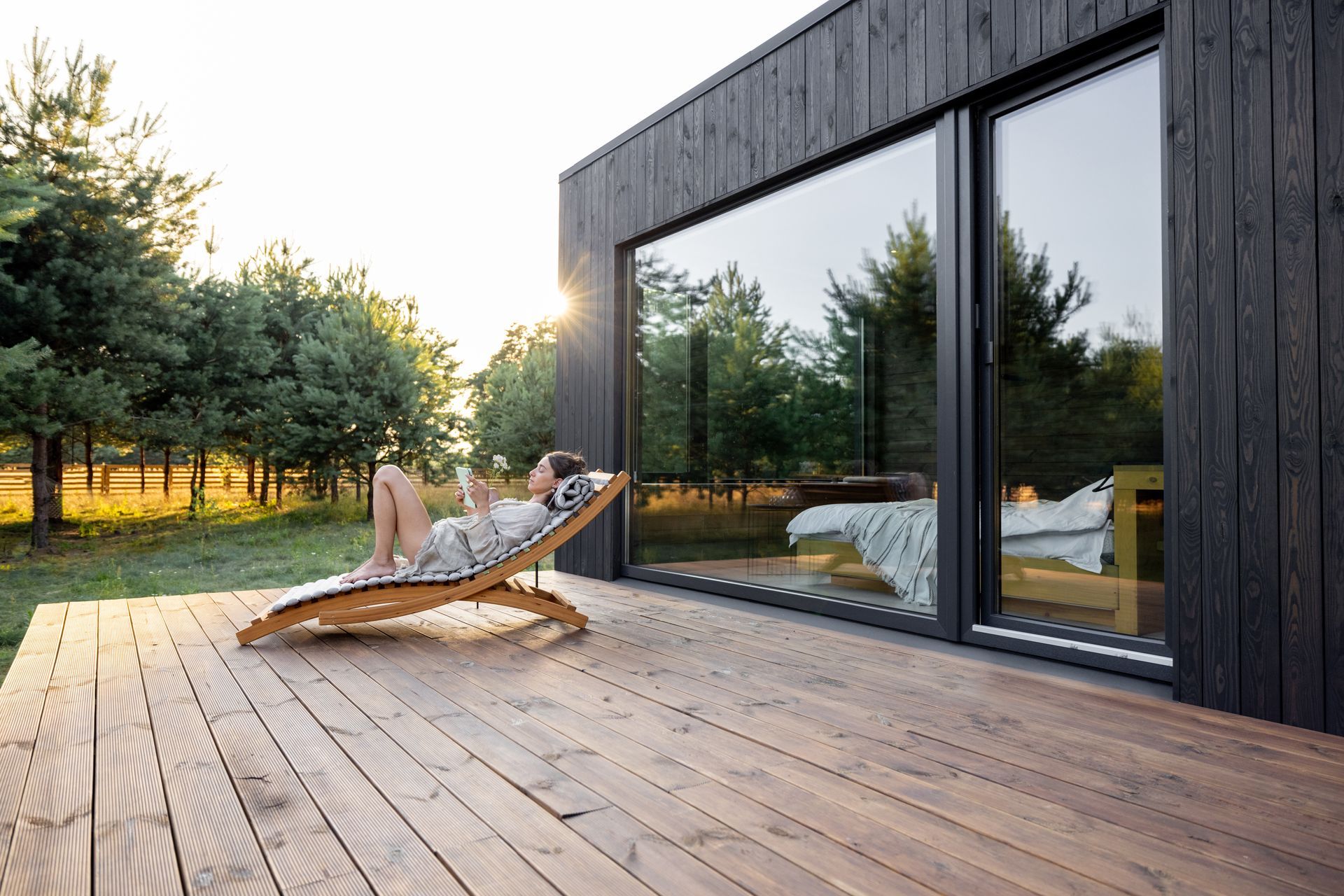 Une personne allongée sur une chaise longue en bois près de grandes baies vitrées dans un chalet moderne.