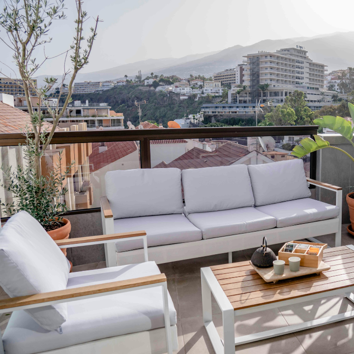 Zona de estar al aire libre con cojines blancos, mesa de madera y vista al paisaje urbano.