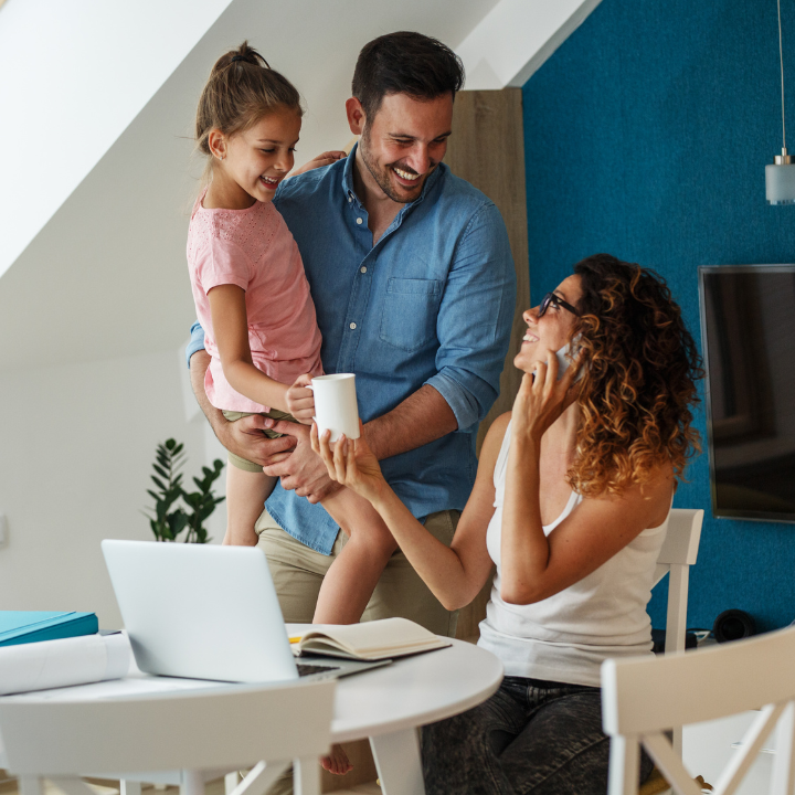 Familia en casa, mujer al teléfono, hombre sosteniendo a un niño, trabajando en la mesa, sonriendo.
