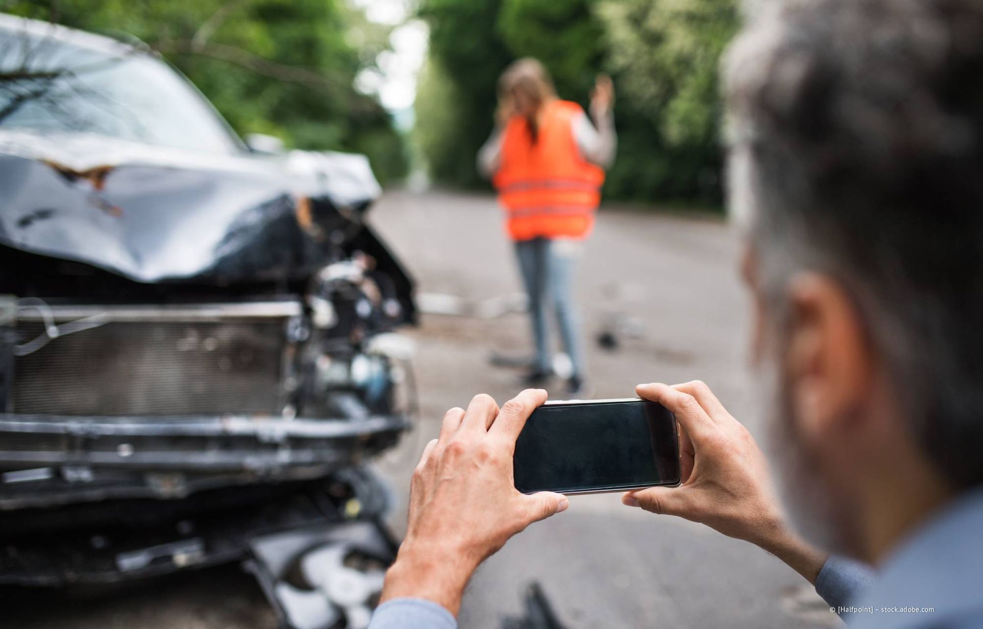 Eine Person fotografiert ein beschädigtes Auto und eine andere Person in einer Warnweste nach einem Autounfall.