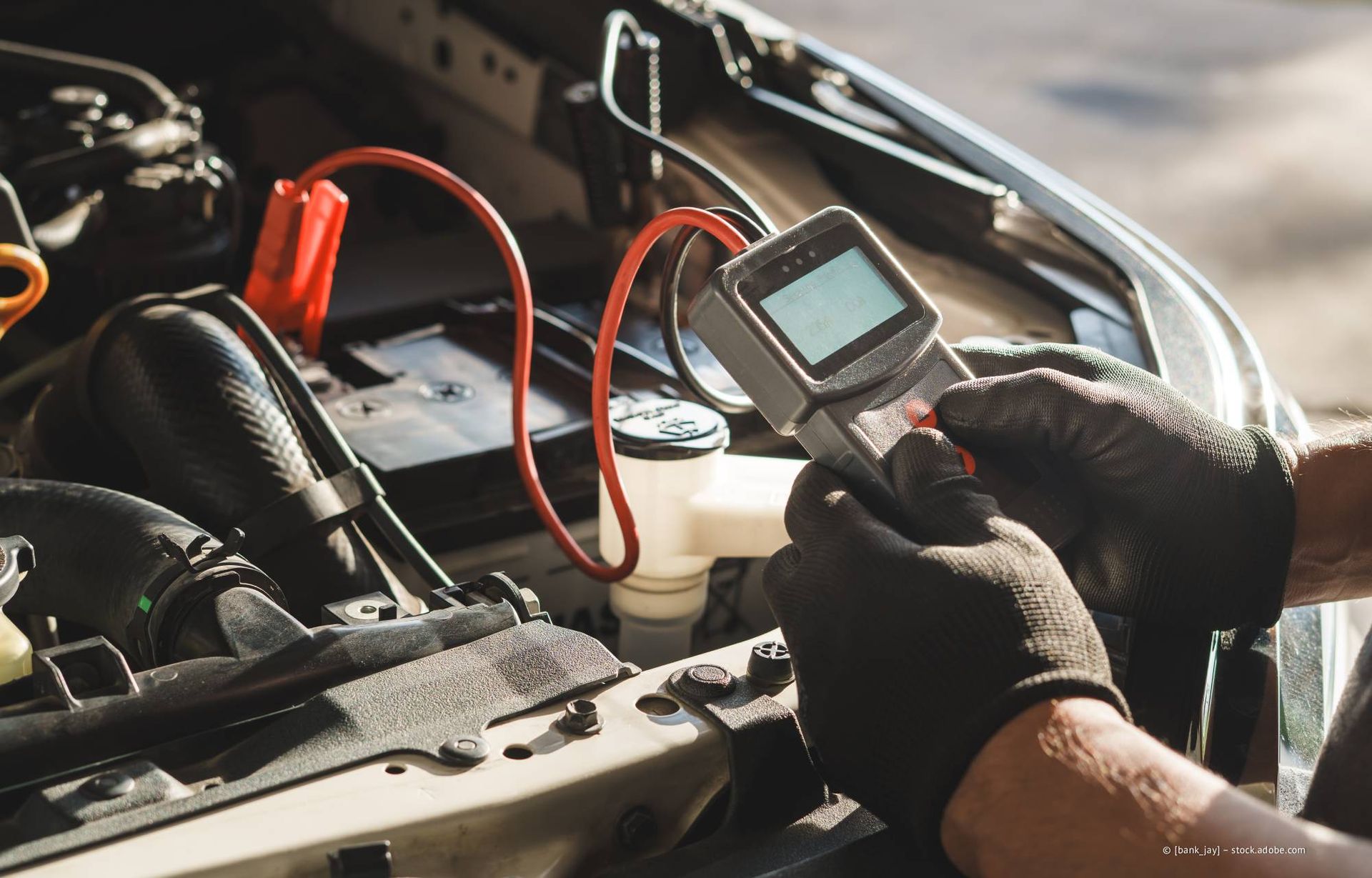 Eine Person mit schwarzen Handschuhen testet im Motorraum mit einem digitalen Multimeter, das an Starthilfekabel angeschlossen ist, eine Autobatterie.