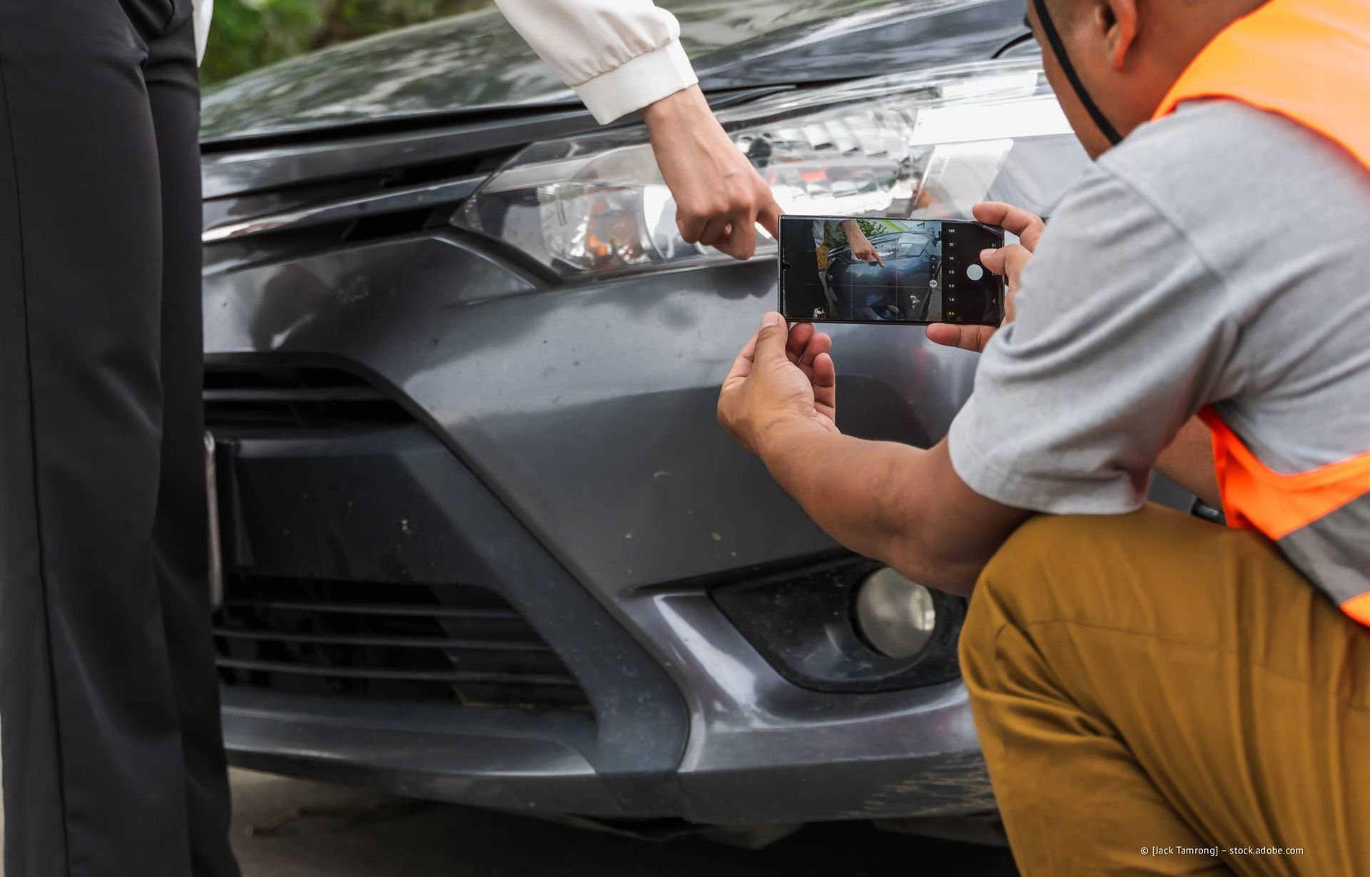 Eine Person fotografiert die beschädigte vordere Stoßstange eines Autos. Eine andere Person zeigt auf den Schaden.