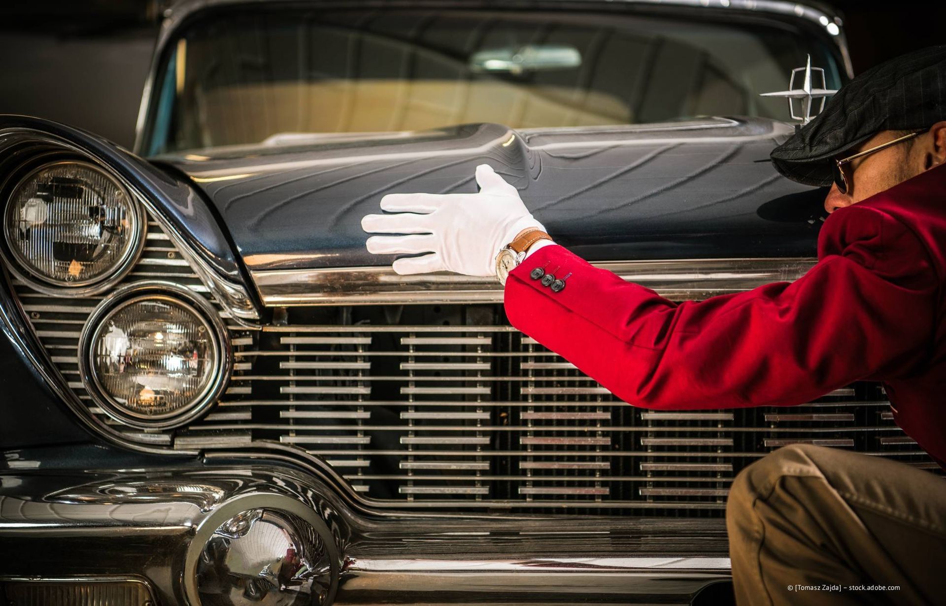Eine Person in roter Jacke und weißen Handschuhen berührt die Motorhaube eines Oldtimers und konzentriert sich dabei auf den Kühlergrill und die Chromteile.