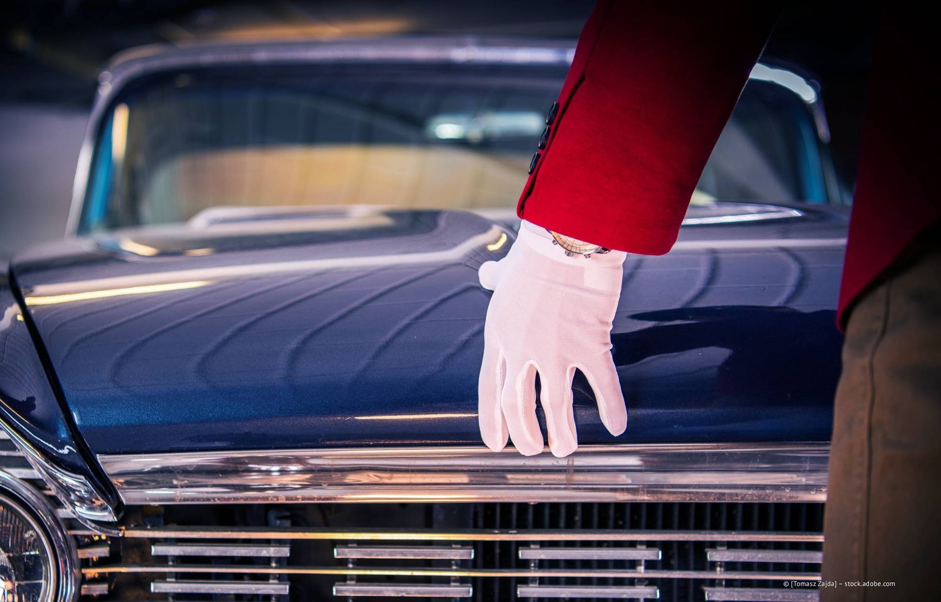 Eine Hand im weißen Handschuh ruht auf der Motorhaube eines klassischen blauen Autos; der rote Jackenärmel ist sichtbar.
