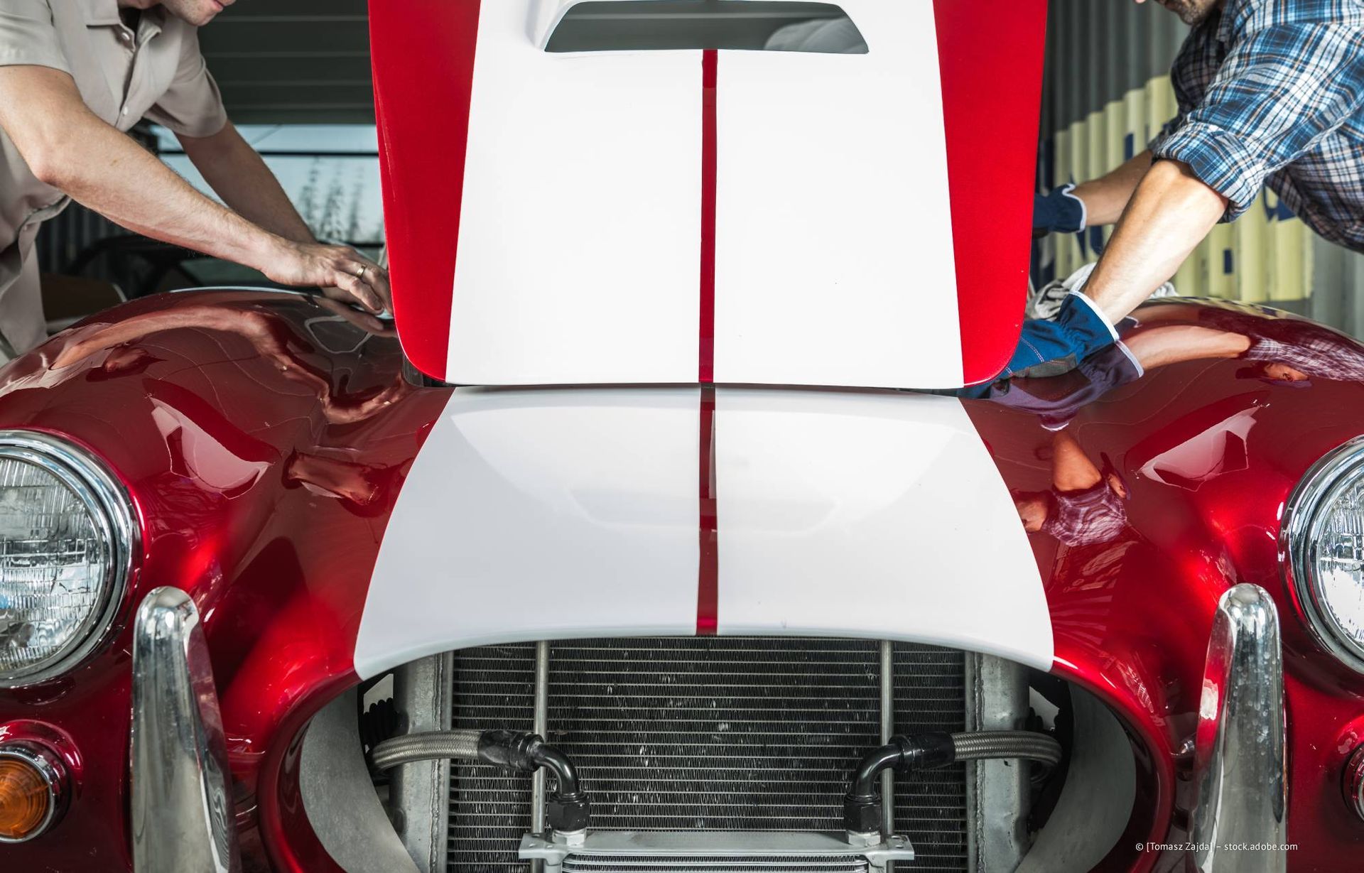 Zwei Personen arbeiten in einer Garage an der geöffneten Motorhaube eines rot-weißen Oldtimers.