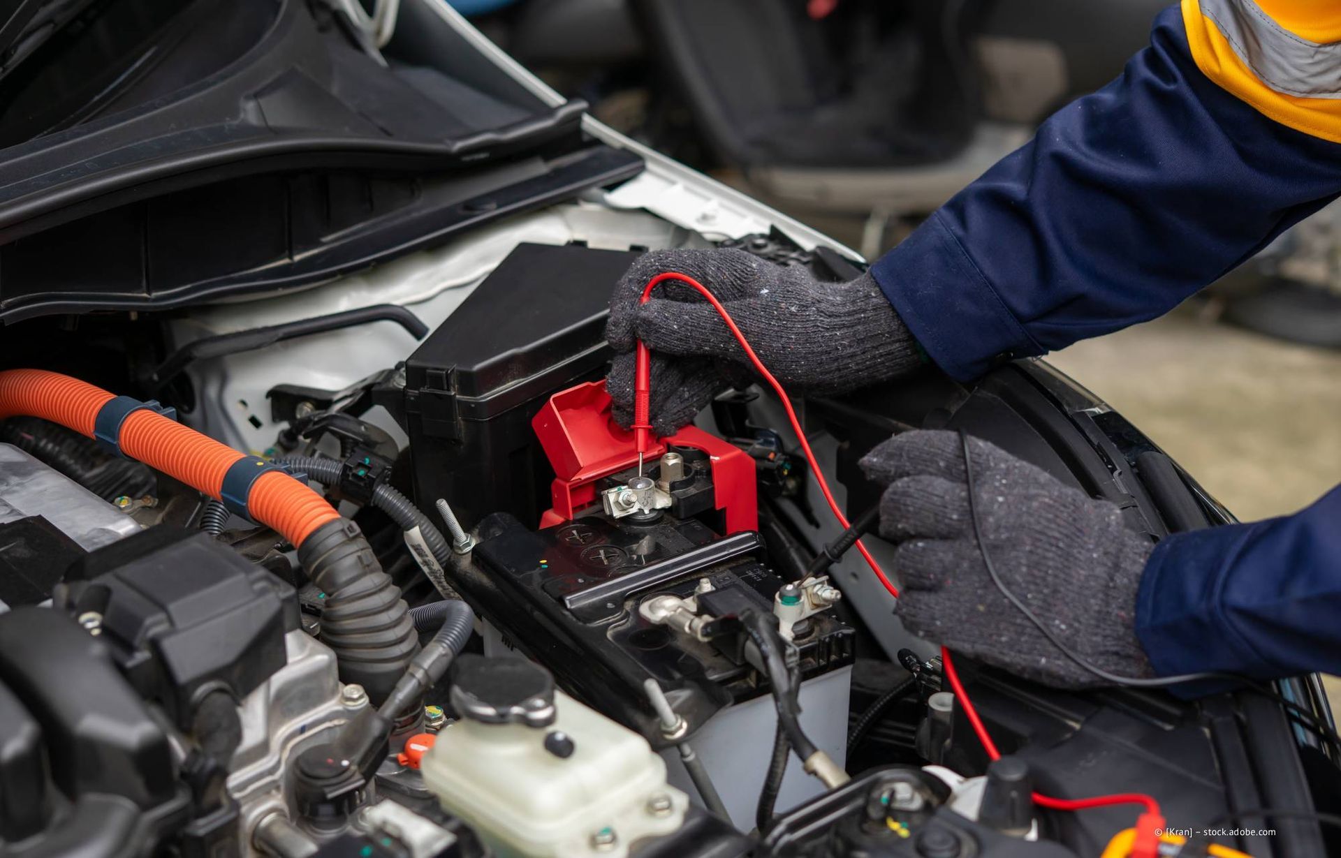 Die behandschuhten Hände eines Mechanikers testen mit einem Multimeter die Autobatterie unter der Motorhaube.