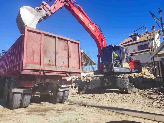 Excavadora roja cargando un camión volquete rojo en un sitio de construcción, con una casa al fondo.