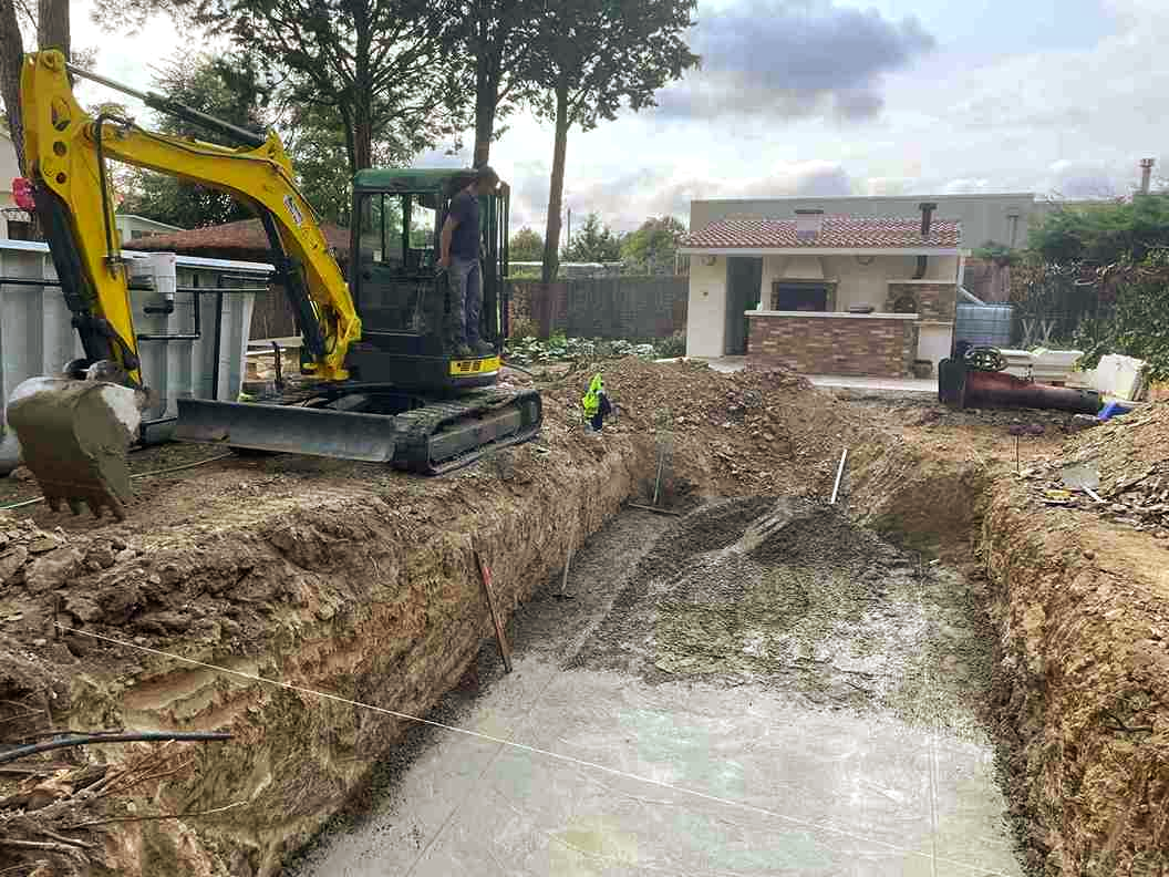Excavadora cavando una zanja para una piscina en un patio trasero; cocina al aire libre visible en el fondo.