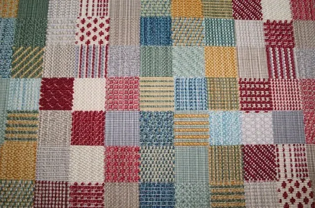 Una colcha o tela de retazos compuesta por pequeños cuadrados de diferentes colores y con distintos patrones texturizados.