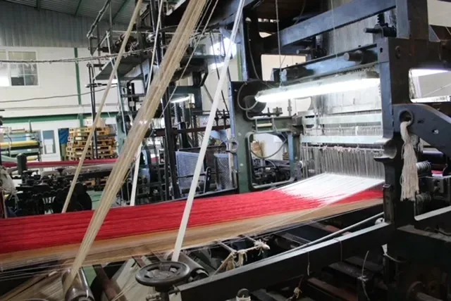 Un telar industrial tejiendo tela roja en el entorno de una fábrica textil.