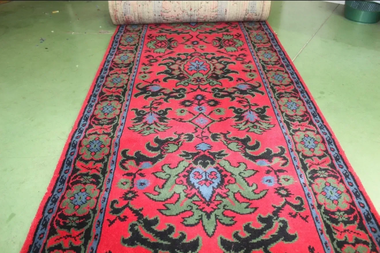 Una alfombra roja con intrincados motivos florales en negro, verde y azul, parcialmente desenrollada sobre un suelo verde.