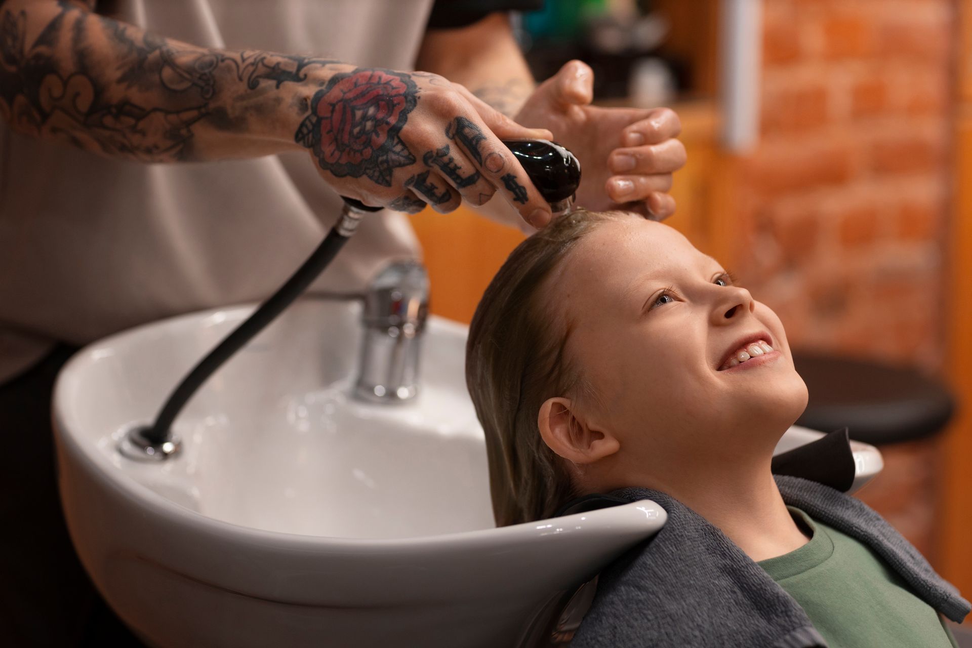 Una estilista tatuada enjuaga el cabello de una clienta sonriente en el lavabo de la peluquería.