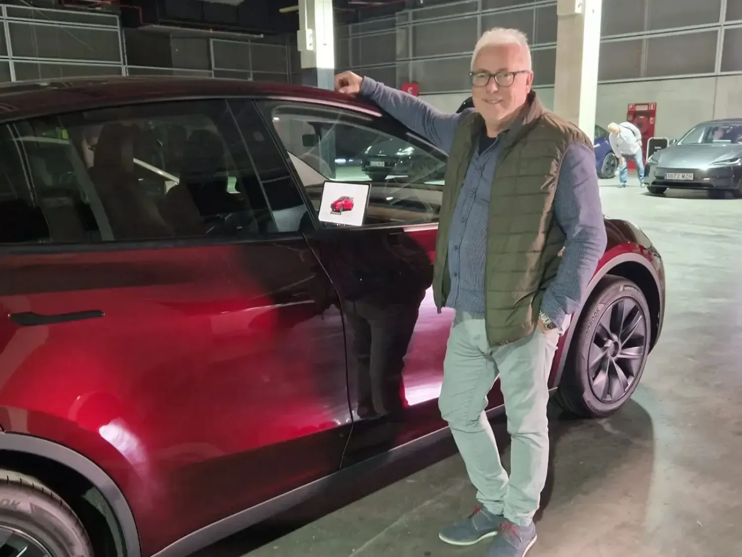 Un Tesla plateado aparcado frente a una terminal de aeropuerto con tres maletas alineadas a su lado.