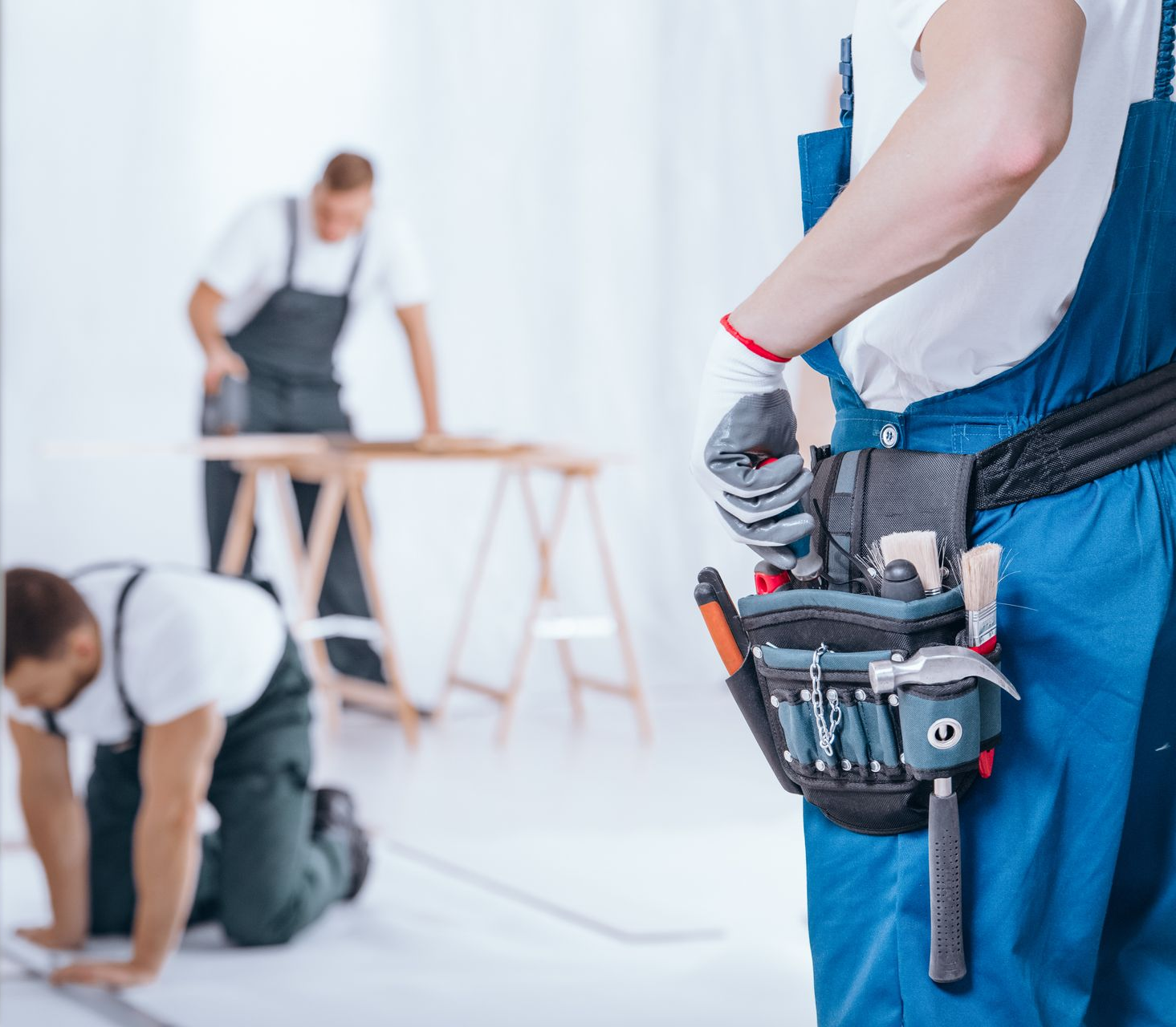 Des ouvriers du bâtiment en salopette bleue travaillent dans une pièce lumineuse. L'un d'eux tient une ceinture porte-outils.