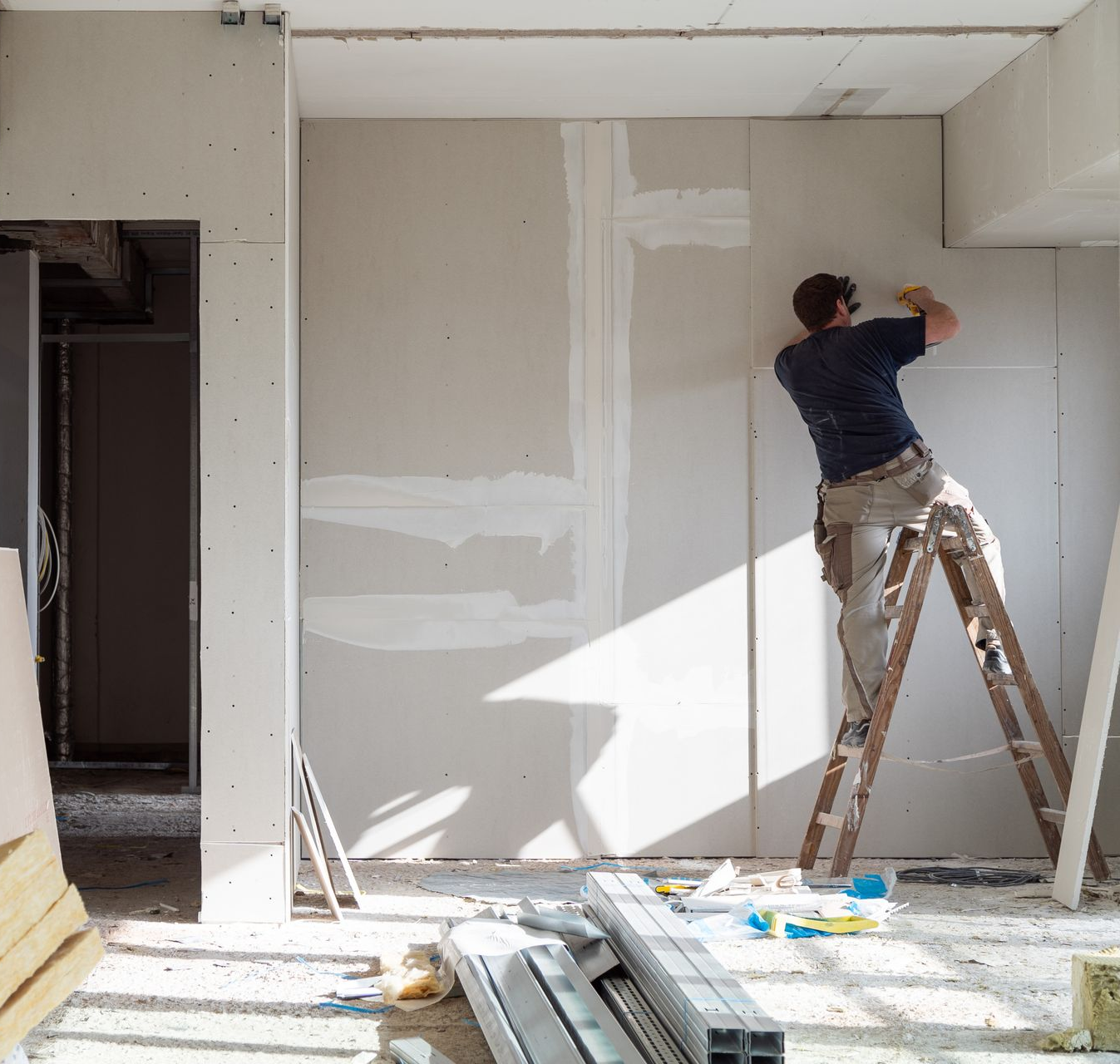 Un homme sur une échelle installe des plaques de plâtre sur un chantier de construction baigné de soleil.