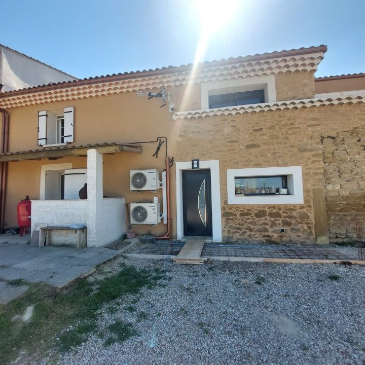 Extérieur d'une maison à deux étages avec des murs en stuc brun clair et en pierre, sous un ciel ensoleillé.