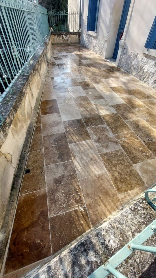 Terrasse en pierre avec dalles rectangulaires brun clair et brun foncé. Rambarde bleue et bâtiment aux volets bleus visibles.