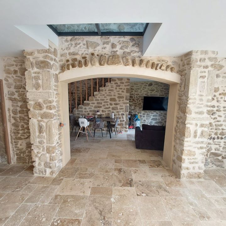 Vue intérieure : passage voûté en pierre menant à une salle à manger et à un escalier ; murs en pierre, sol carrelé, plafond en verre.