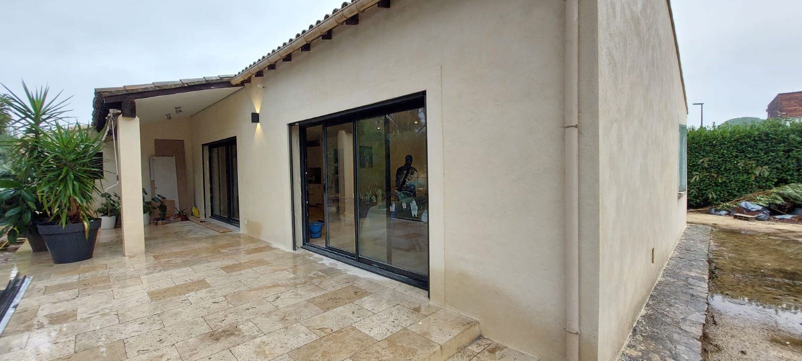 Vue extérieure d'une maison beige avec un patio couvert et des portes coulissantes en verre, par temps de pluie.