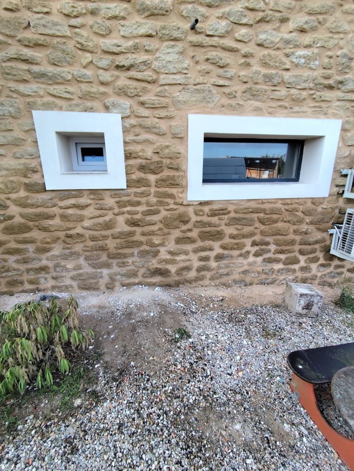 Mur en pierre percé de deux fenêtres ; l'une carrée, l'autre rectangulaire, toutes deux à encadrement blanc.