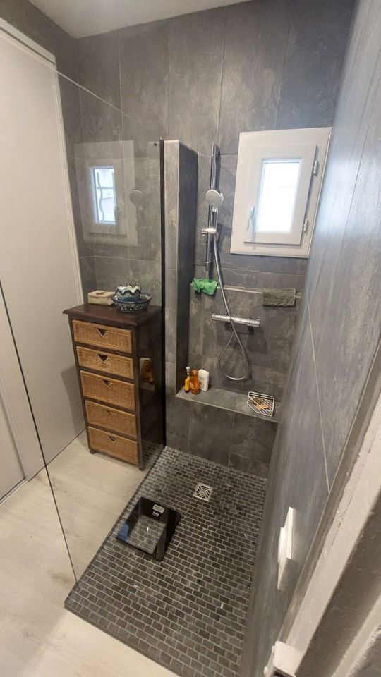 Salle de bains avec douche carrelée grise et tiroirs en bois près de la fenêtre.