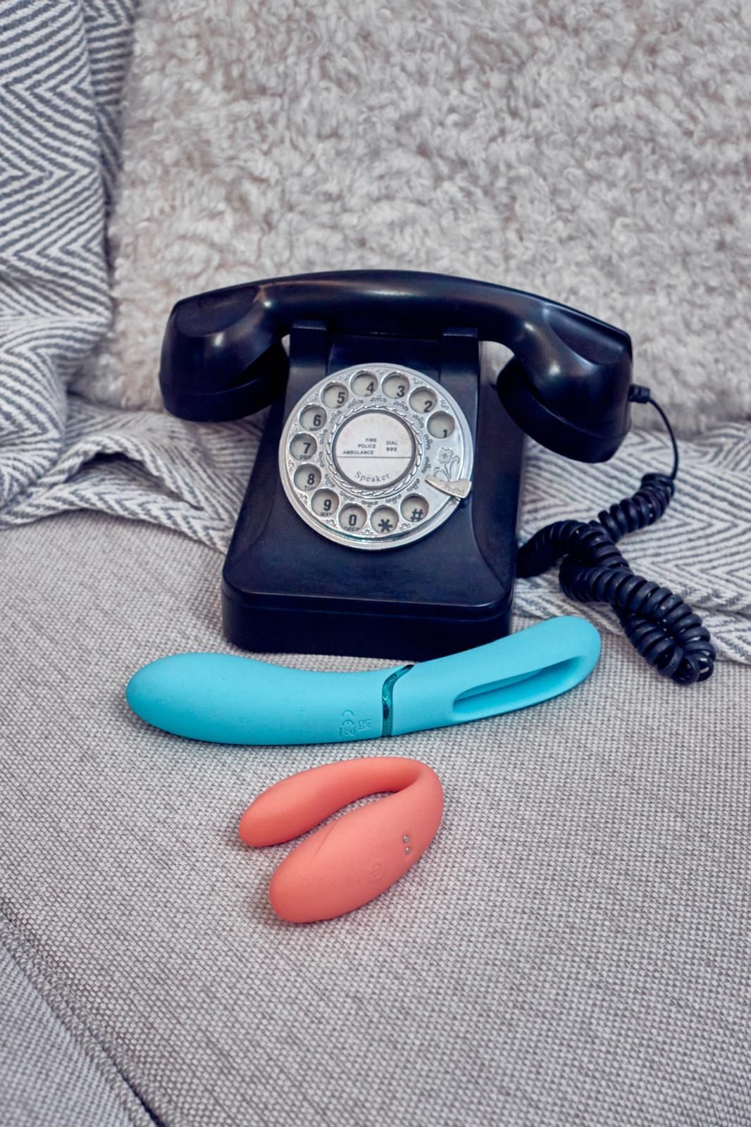 Una mujer con gafas señala juguetes sexuales sobre una mesa, sosteniendo una varita mágica. En un entorno con poca luz y vegetación.