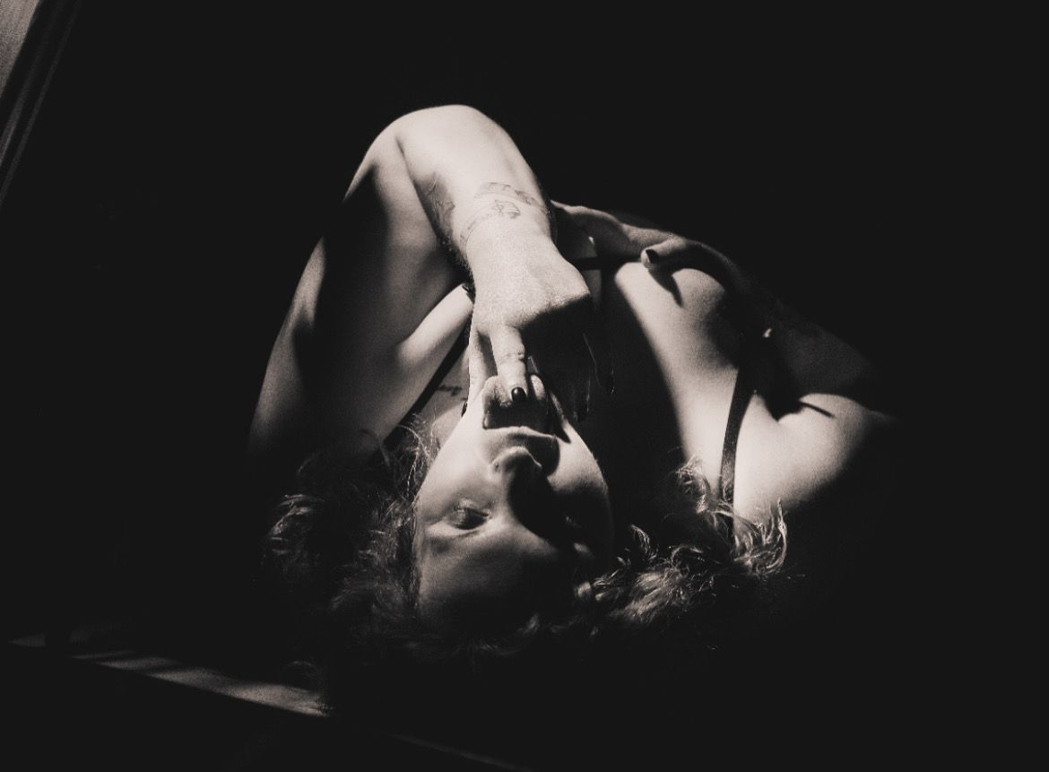 La mujer se reclina, con la cabeza inclinada hacia arriba y el brazo sobre la cara, en un entorno oscuro con foco.